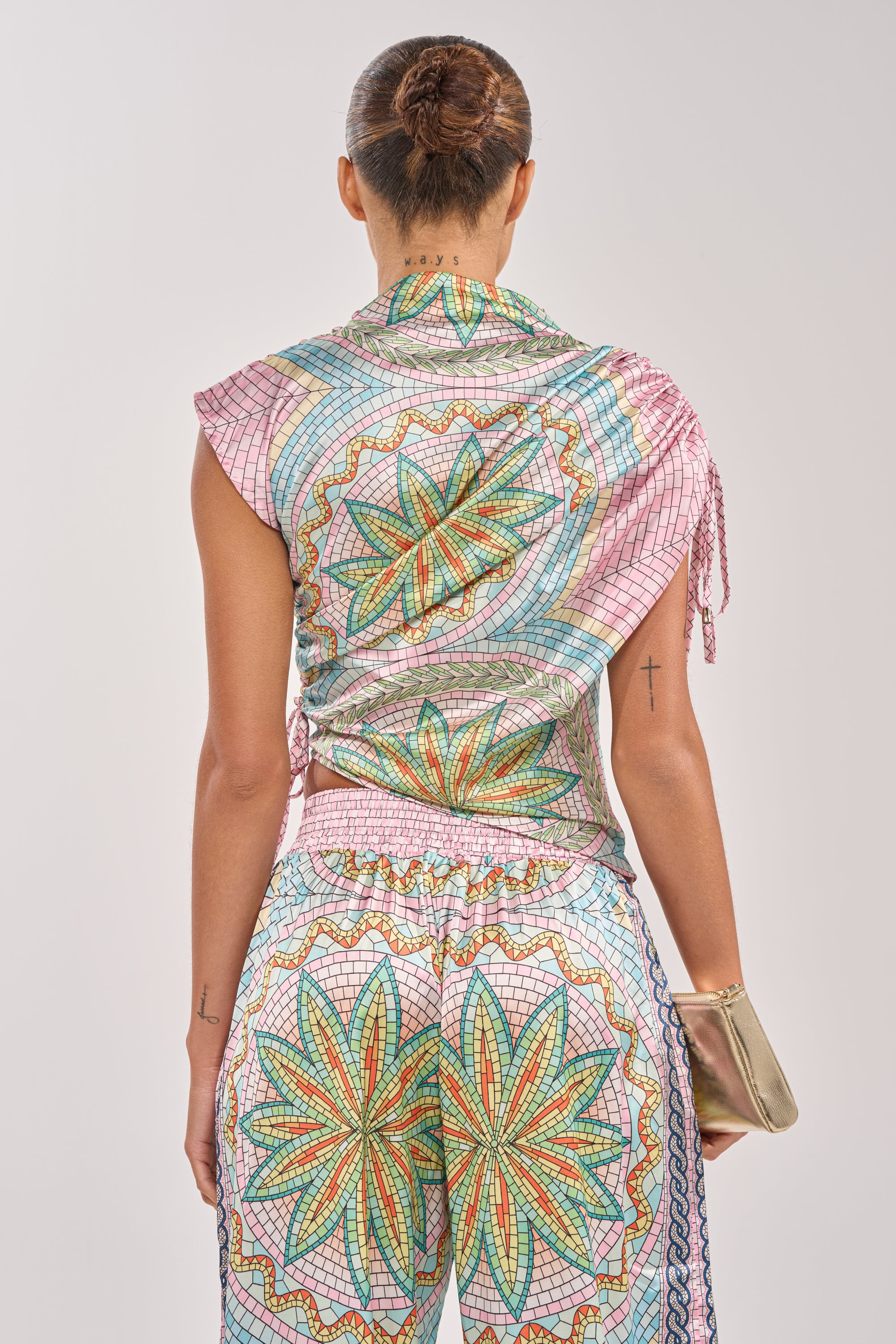 A woman with her hair in a bun, viewed from behind, wears the ALL DIFFERENT WAYS SATIN BLOUSE in pink mosaic with green, red, and yellow floral designs. She holds a gold clutch and has tattoos on her neck and arms. 4