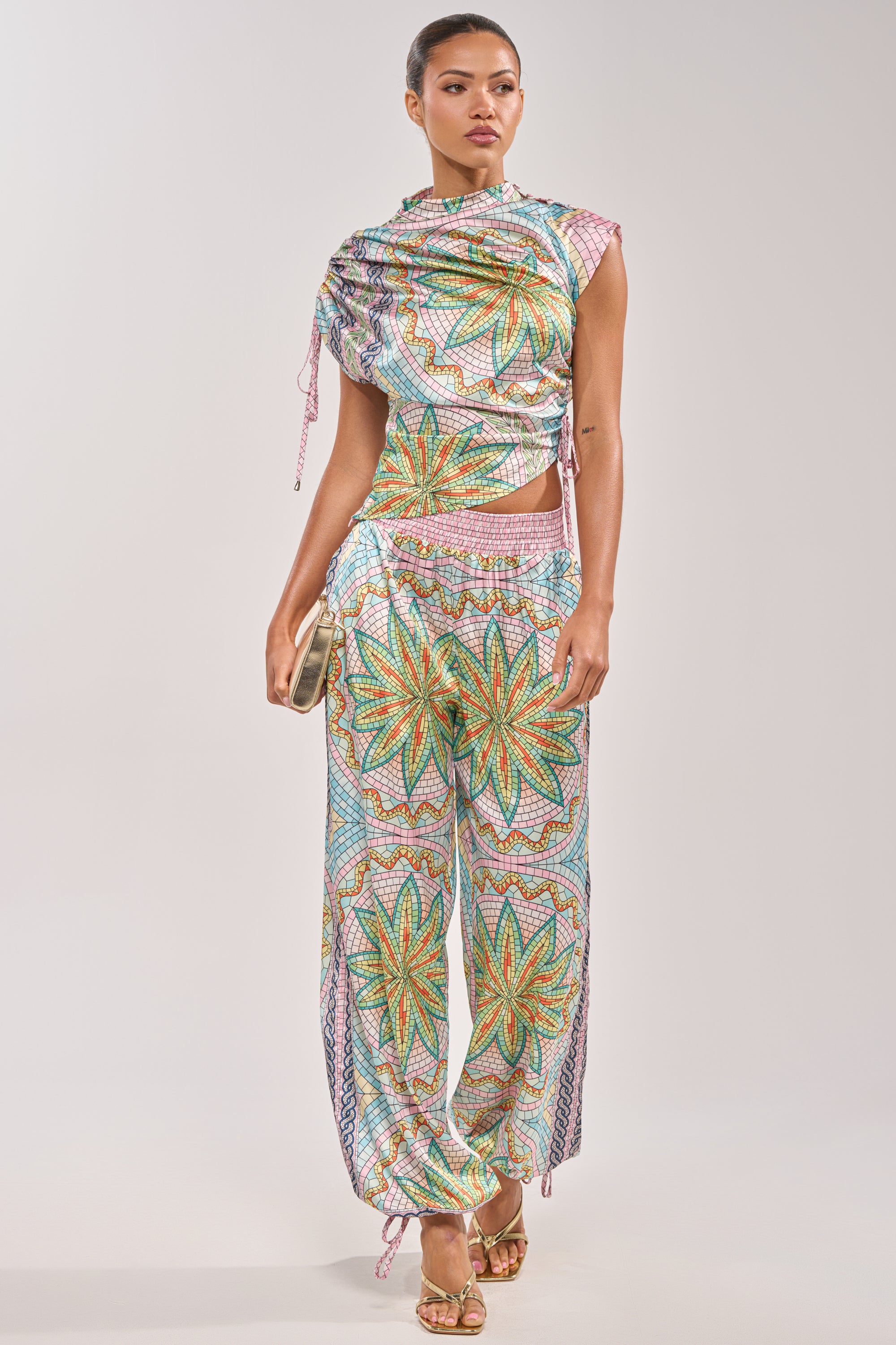 A woman wears the ALL DIFFERENT WAYS SATIN BLOUSE, a vibrant draped top with cutouts and star patterns, paired with matching loose pants. She accessorizes with a gold clutch and strappy heels. 5