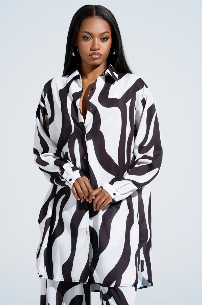 Wearing the AKIRA Label's LET IT GO BUTTON DOWN TOP with a bold black and white abstract swirl, a model poses calmly against a light background with long, straight hair.