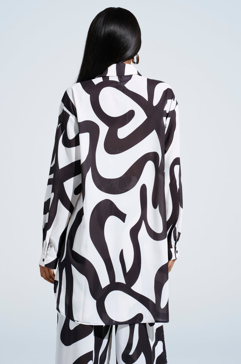 A long-haired person in the AKIRA Label LET IT GO BUTTON DOWN TOP with matching abstract swirl pants stands back to the camera against a light background.