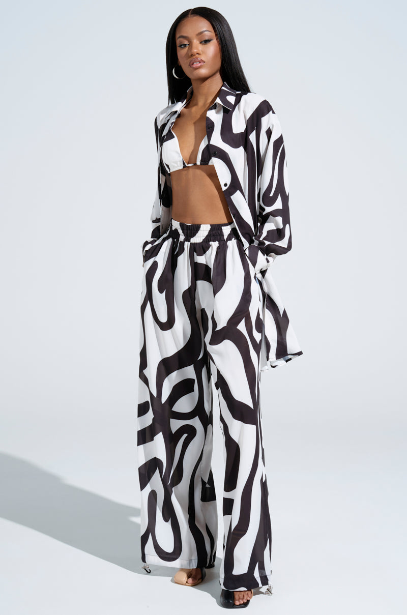 A person poses confidently in an AKIRA Label outfit featuring the black and white abstract swirl LET IT GO BUTTON DOWN TOP, bralette, and wide-leg pants. They stand on a light background with one hand in their pocket and the other on their hip.