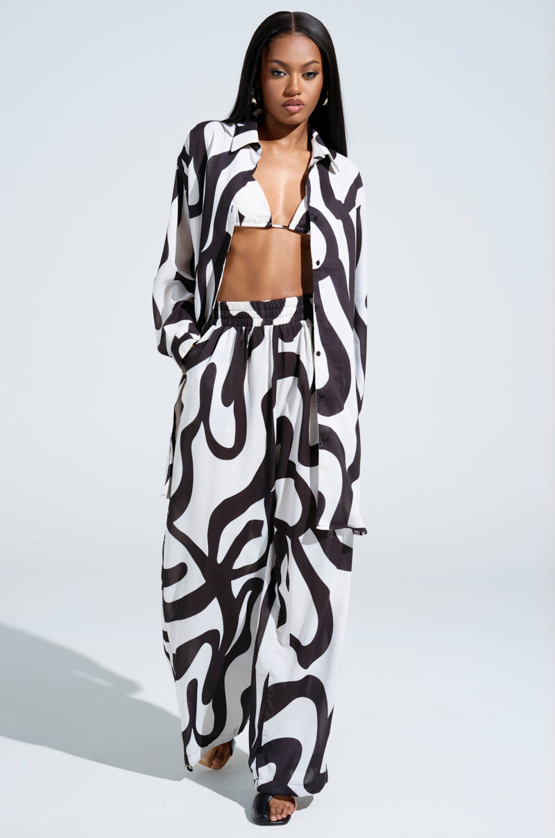 A woman confidently poses in the Let It Go Button Down Top and matching flowy pants, featuring an abstract swirl pattern over a black crop top, while standing against a plain backdrop and looking directly at the camera.
