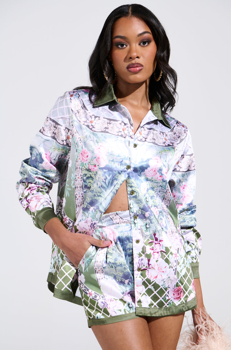 A woman poses confidently in a floral-patterned outfit featuring the Flower Party Satin Button Up Shirt with a unique cutout and matching shorts. She has her hand in one pocket, wears hoop earrings, with her long hair styled loosely. The background is light gray.