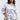 A woman poses confidently in a floral-patterned outfit featuring the Flower Party Satin Button Up Shirt with a unique cutout and matching shorts. She has her hand in one pocket, wears hoop earrings, with her long hair styled loosely. The background is light gray.