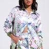 A woman poses confidently in a floral-patterned outfit featuring the Flower Party Satin Button Up Shirt with a unique cutout and matching shorts. She has her hand in one pocket, wears hoop earrings, with her long hair styled loosely. The background is light gray.