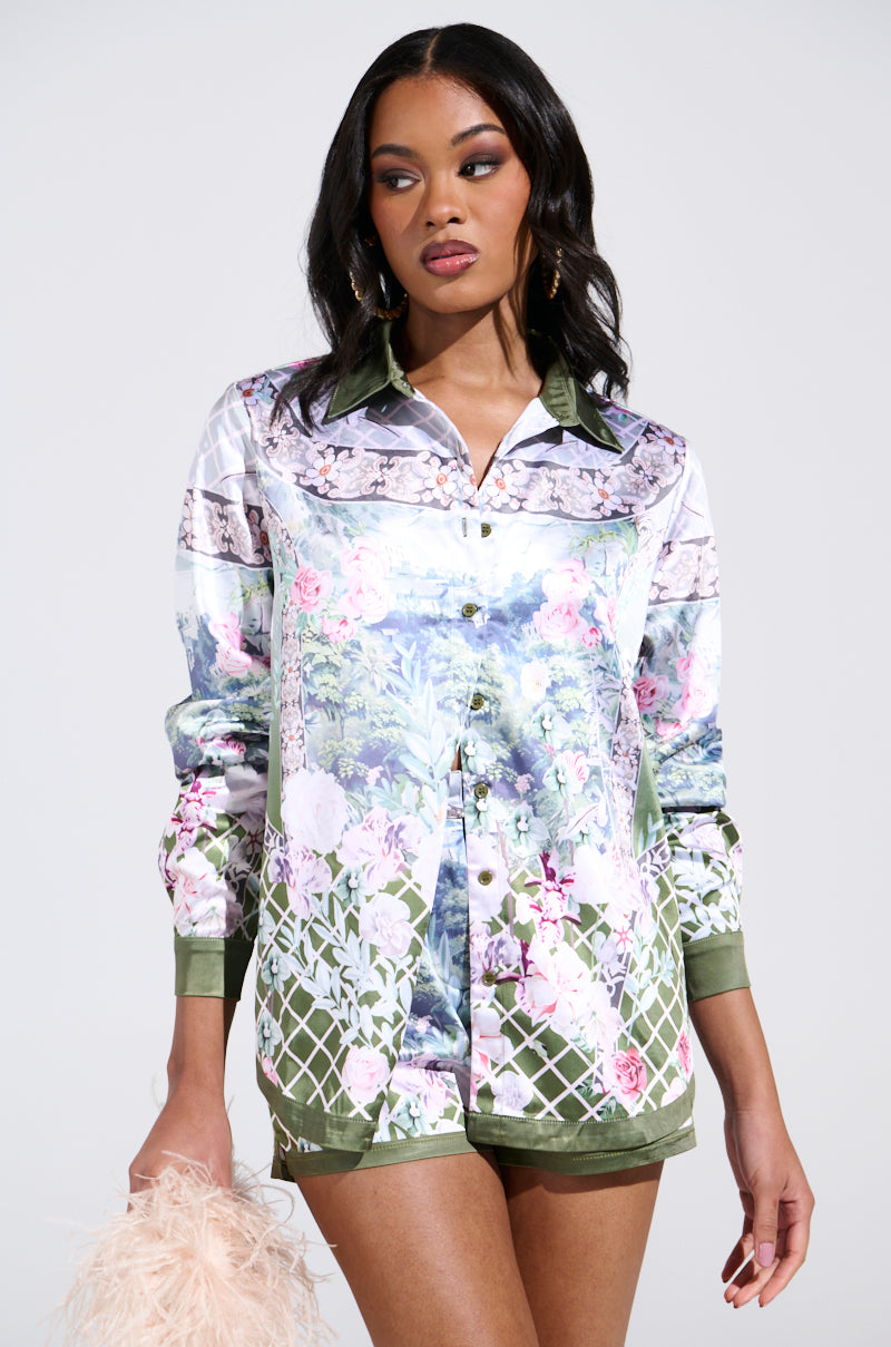 A woman in the FLOWER PARTY SATIN BUTTON UP SHIRT with a floral pattern and olive green accents holds a fuzzy light pink object. Her long dark hair cascades over her shoulders against the plain backdrop.
