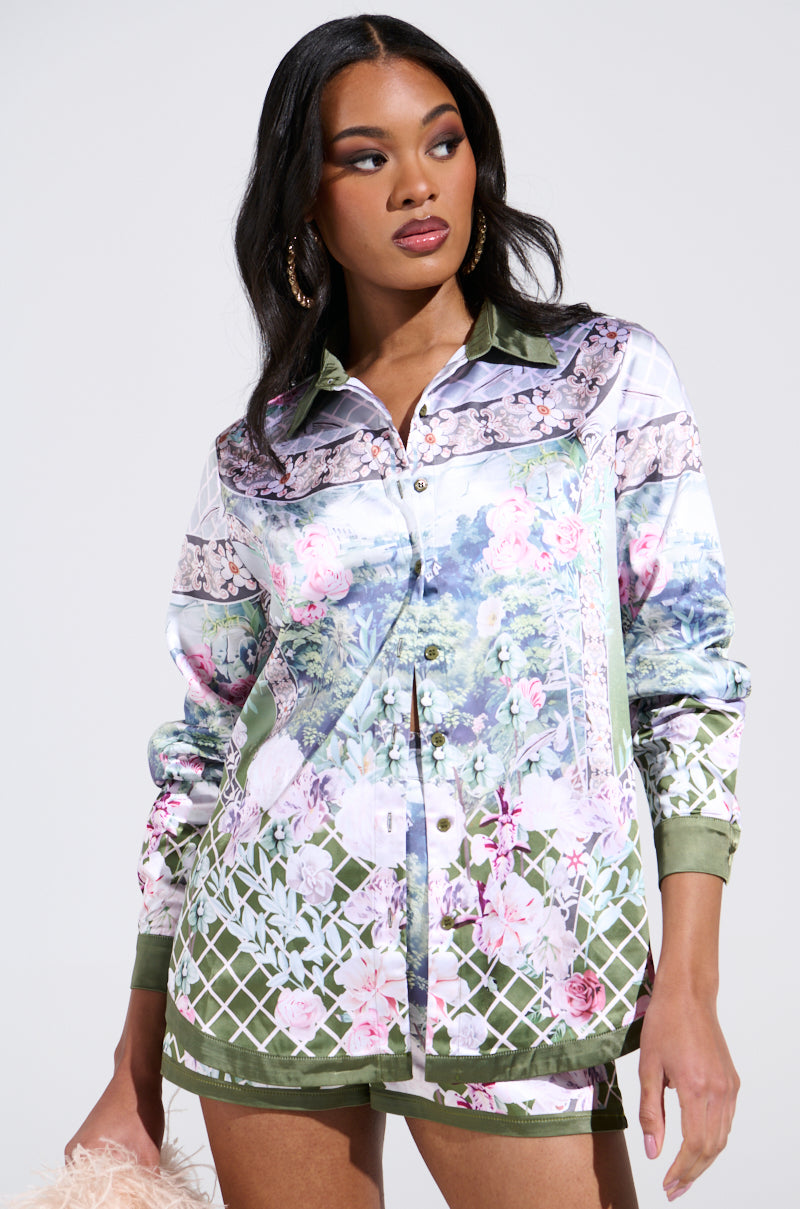 A woman poses against a plain background in the FLOWER PARTY SATIN BUTTON UP SHIRT and matching floral shorts. Her hair flows in loose waves as she holds a fluffy pink accessory, exuding effortless elegance.