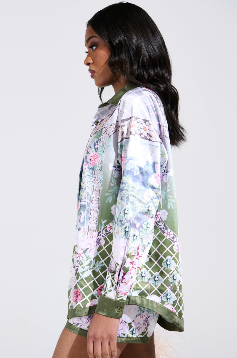 A woman with long, dark hair is seen in profile, wearing the FLOWER PARTY SATIN BUTTON UP SHIRT adorned with vibrant green, pink, and blue flowers, set against a plain background.