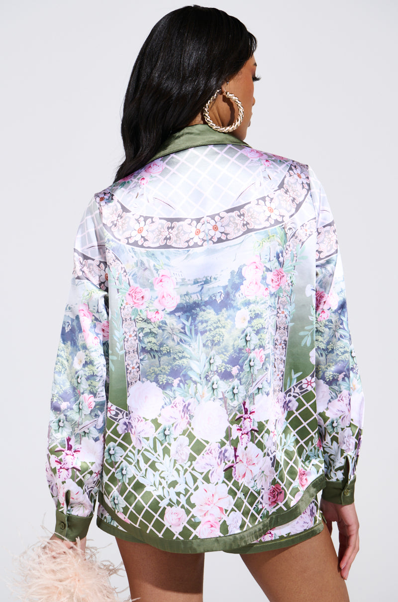 A person with long dark hair models the FLOWER PARTY SATIN BUTTON UP SHIRT, featuring a silky floral pattern with scenic print and green accents. Large hoop earrings complement their look as they hold a fluffy pink accessory, exuding elegant style with their back to the camera.