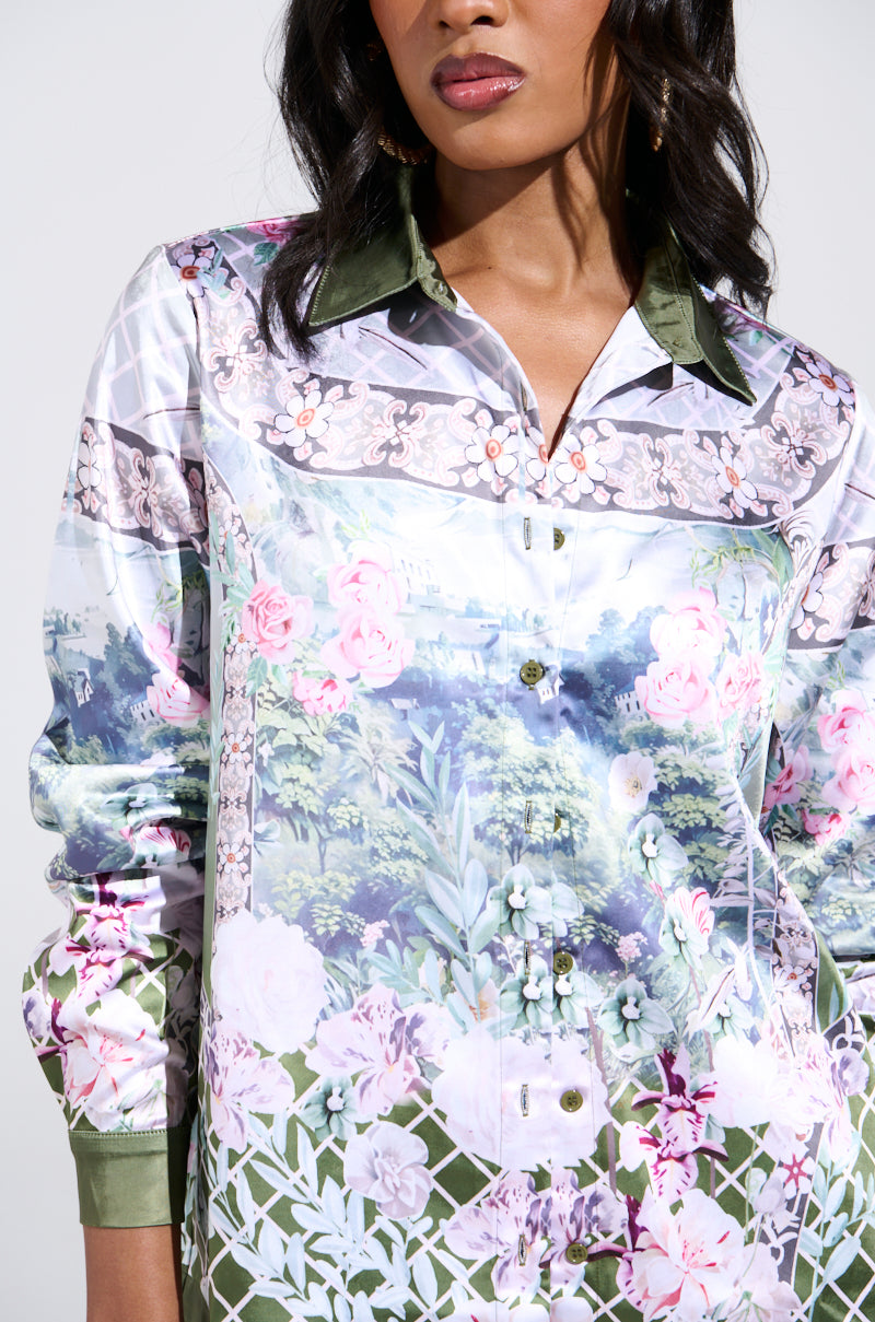 A woman wears the FLOWER PARTY SATIN BUTTON UP SHIRT, featuring a colorful floral pattern with a green collar and pink, green, and white flower and foliage imagery against a solid backdrop. Only her upper body is visible.