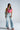 A woman wears the LOVE LOVE LOVE BLOUSE, light blue high-waisted jeans with side detailing, pink heeled boots, and yellow-tinted sunglasses, standing against a plain background.