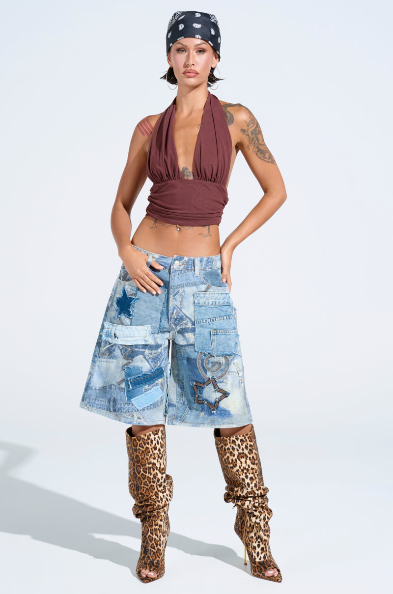 Sporting a PAXTON Sexy Ruched Plunge Mesh Halter Top, patchwork denim culottes, and leopard print heeled boots, the individual stands confidently. Tattoos adorn their arms as they pose with a hand on their waist against a light background, and a bandana adds flair to their bold ensemble.