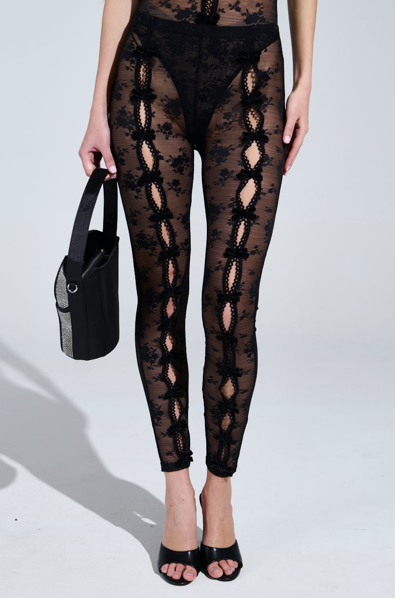 A person models the YOU WILL SEE LACE LEGGING, black with floral lace patterns and front cut-outs. They carry a black handbag and wear black open-toe heels featuring velvet bow appliques, against a plain white background.