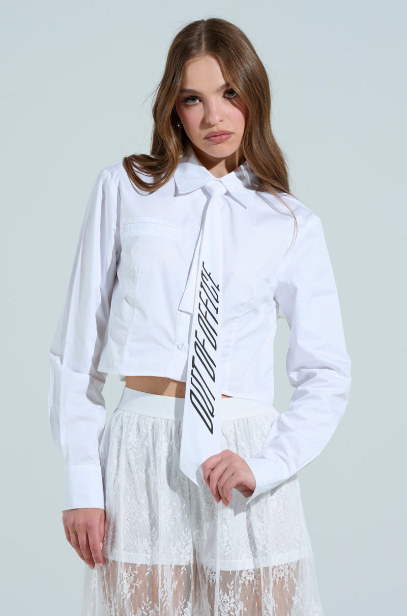 A woman dons the OOO BUTTON DOWN TOP featuring a bold "OUT OF OFFICE" tie, stylishly paired with a white lace skirt. The crisp poplin fabric contrasts elegantly against a light gray background, embodying an effortlessly chic style.