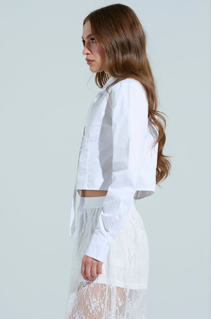A woman with long brown hair is seen in profile, wearing an OOO BUTTON DOWN TOP and a sheer lace skirt against a simple gray background. She is looking forward with a neutral expression.