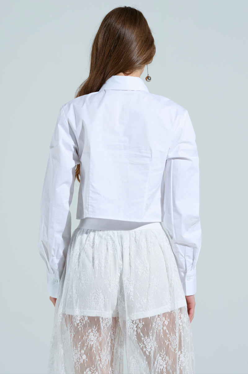 A person with long brown hair is seen from behind wearing the OOO BUTTON DOWN TOP, a crisp white poplin shirt, paired with a delicate lace skirt. The plain, light-colored background gives an "out of office" vibe.