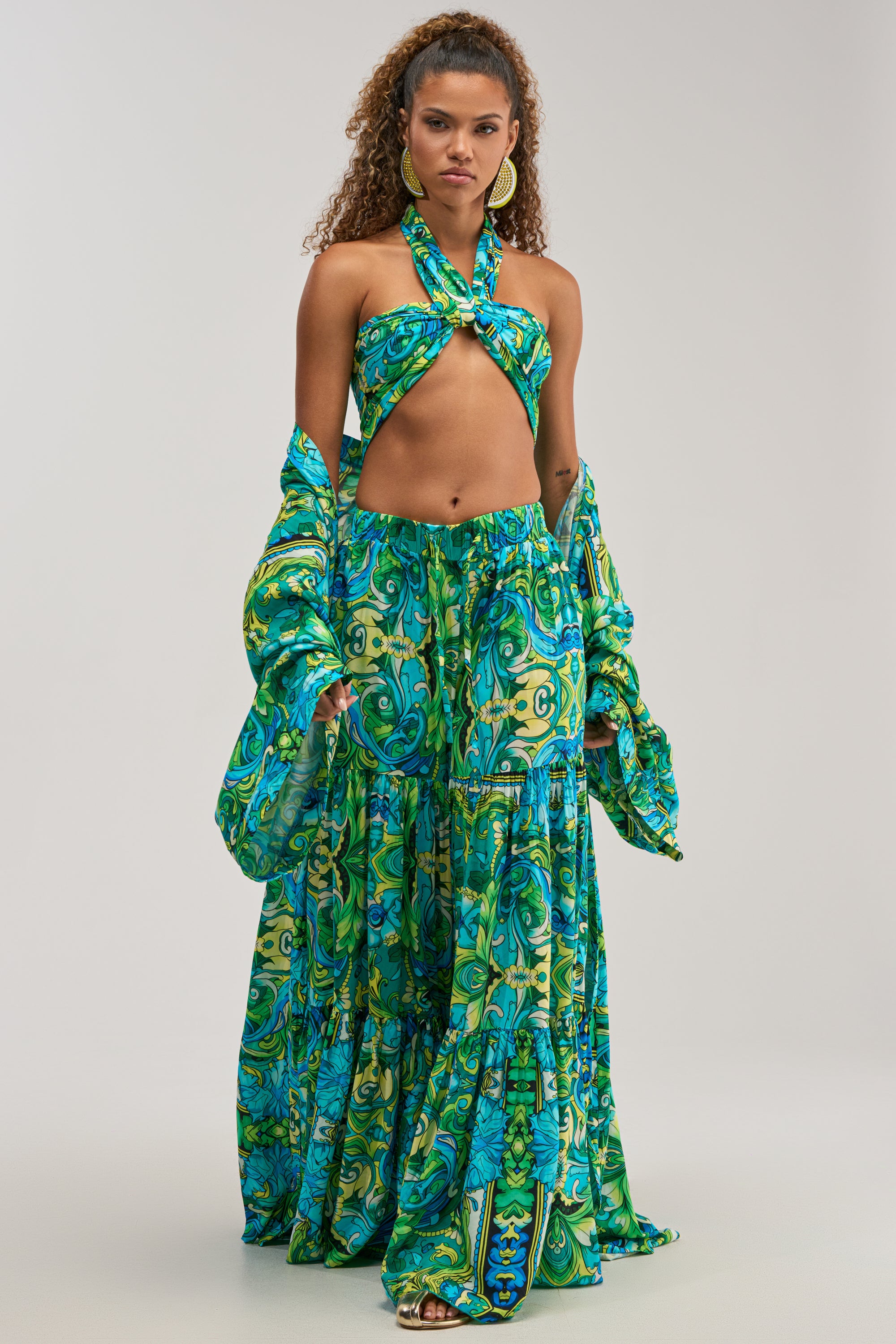 A woman showcases summer style in the INTO THE GARDEN PALAZZO PANT IN GREEN MULTI with a vibrant halter top and matching jacket. Her curly hair and bold earrings complete the look against a light background.