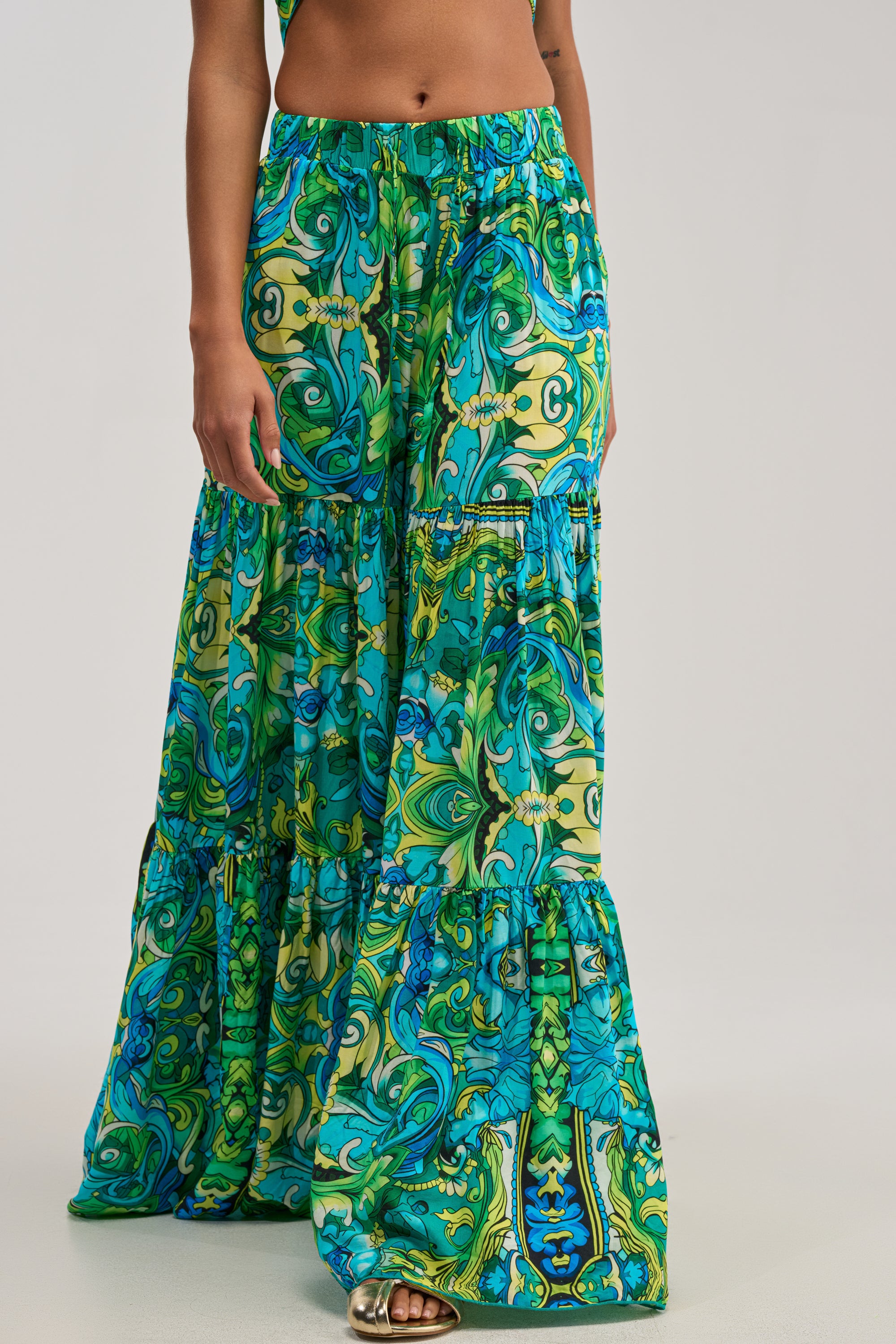 Wearing the INTO THE GARDEN PALAZZO PANT IN GREEN MULTI—vibrant, wide-leg pants with a green and blue botanical print and tiered design—paired with gold sandals, this look is an effortless example of summer styling. The upper body is not fully visible.