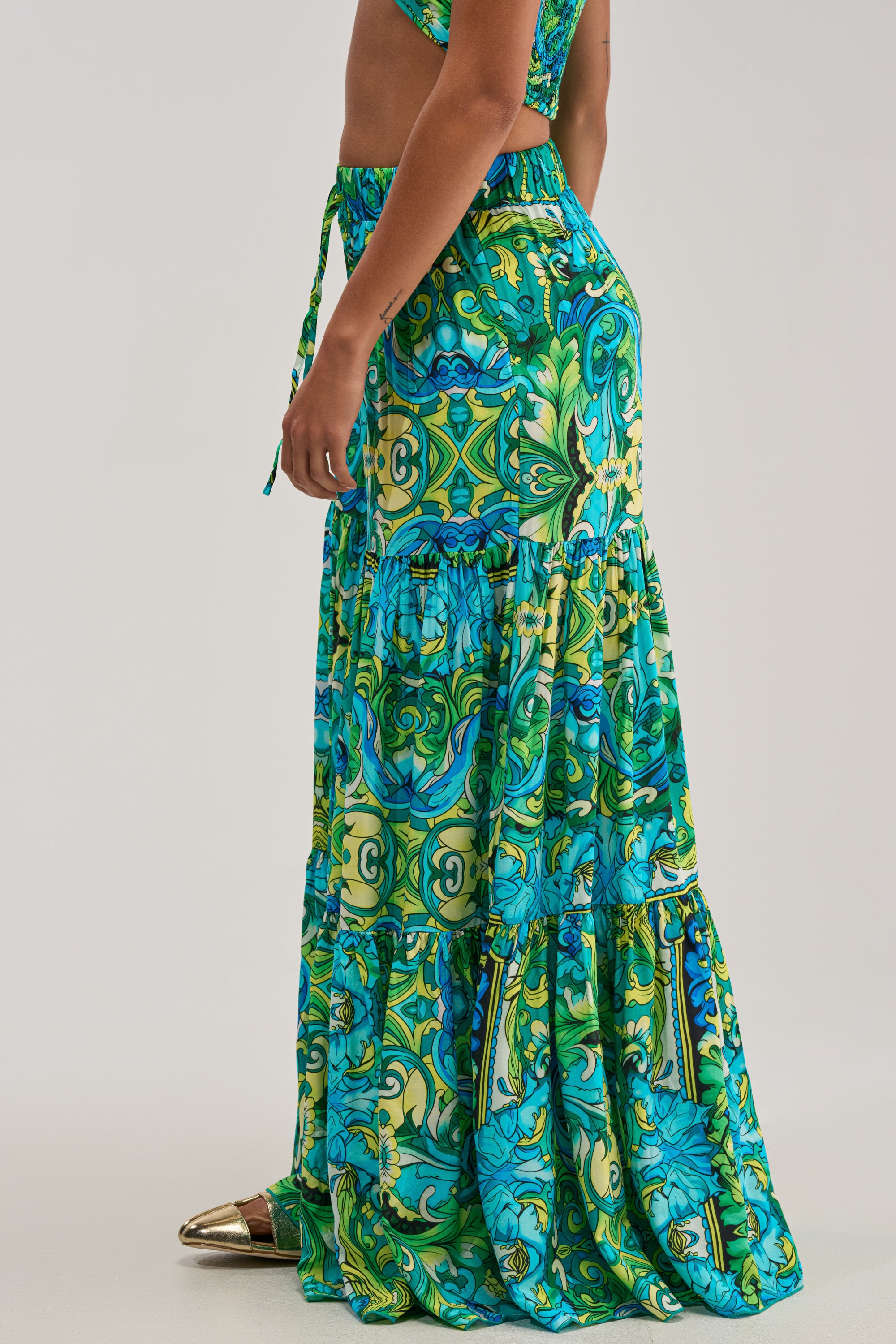 A person stands sideways, showcasing the lower half of their body in the INTO THE GARDEN PALAZZO PANT IN GREEN MULTI, featuring a vibrant green, blue, and yellow pattern and a wide-leg silhouette. Complementing the effortless summer look are gold pointed-toe shoes.