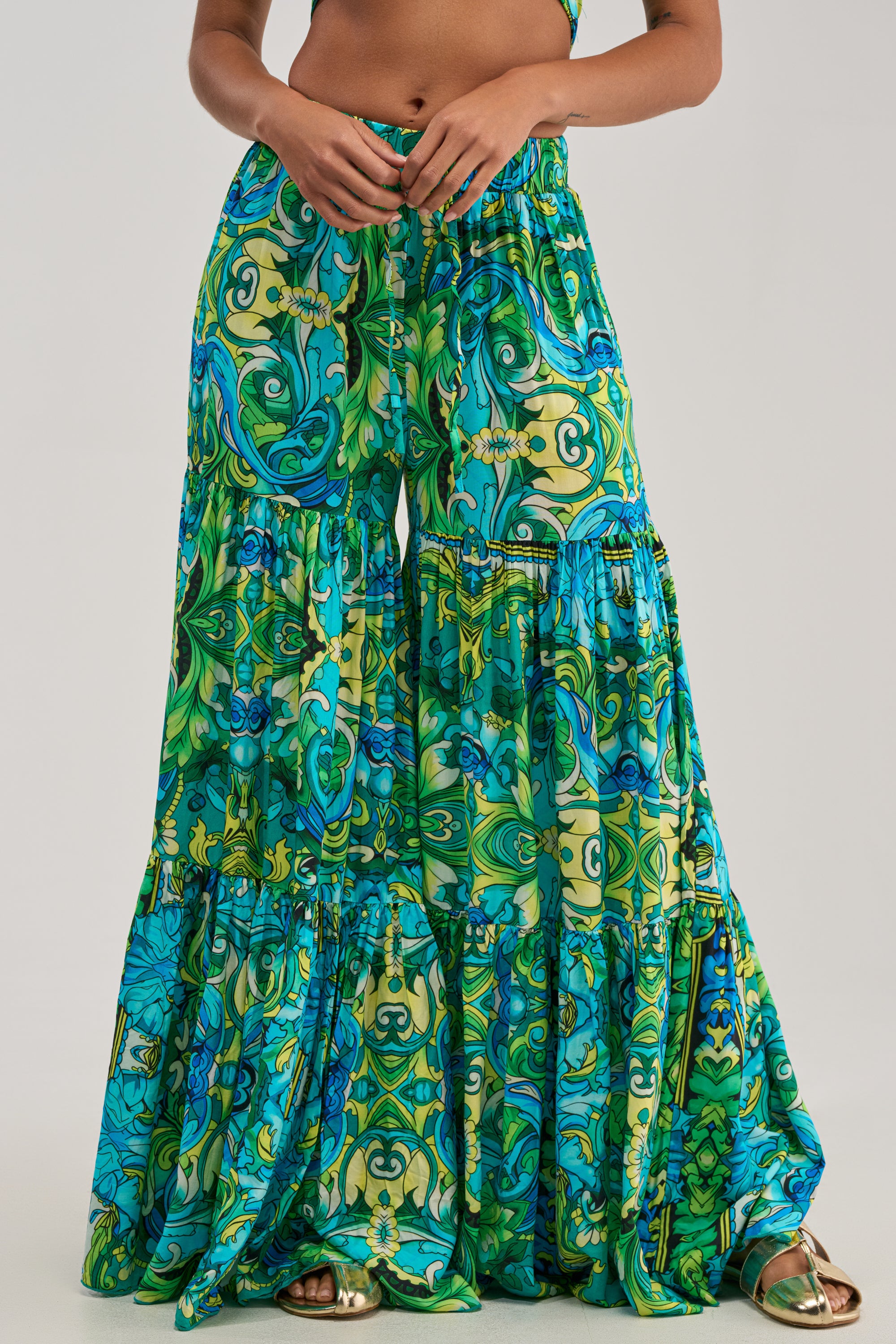 A person showcases the INTO THE GARDEN PALAZZO PANT IN GREEN MULTI, featuring a vibrant green, blue, and yellow floral and geometric print. The flowy, tiered wide-leg silhouette is reminiscent of a botanical palazzo style—perfect for summer styling. Only the lower torso and legs are visible, emphasizing the relaxed fit of these statement pants.