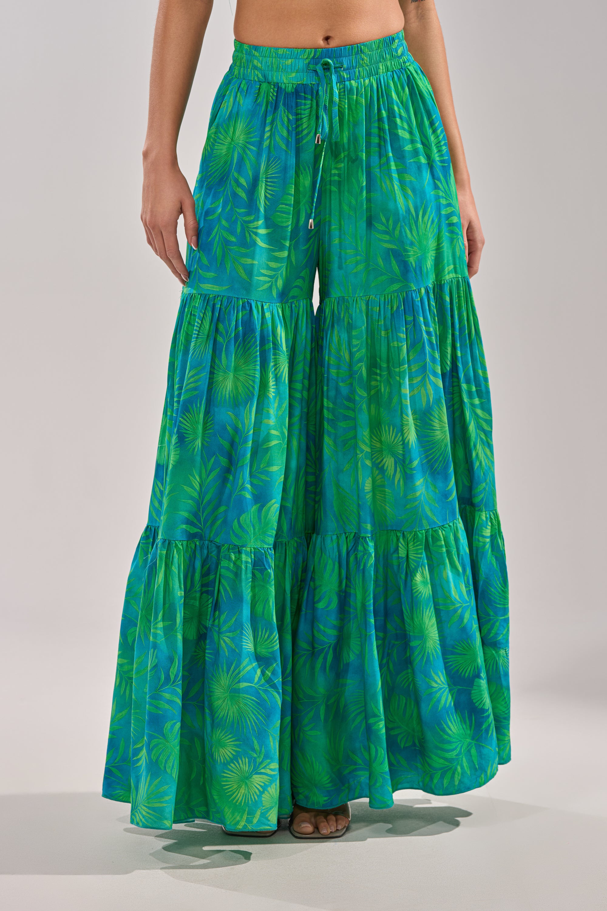 A person stands barefoot on a neutral background wearing the INTO THE GARDEN PALAZZO PANT IN GREEN, featuring wide legs, green and blue tropical leaf print, a drawstring waist, and flowy ruffled tiers for an effortlessly stylish look. 3
