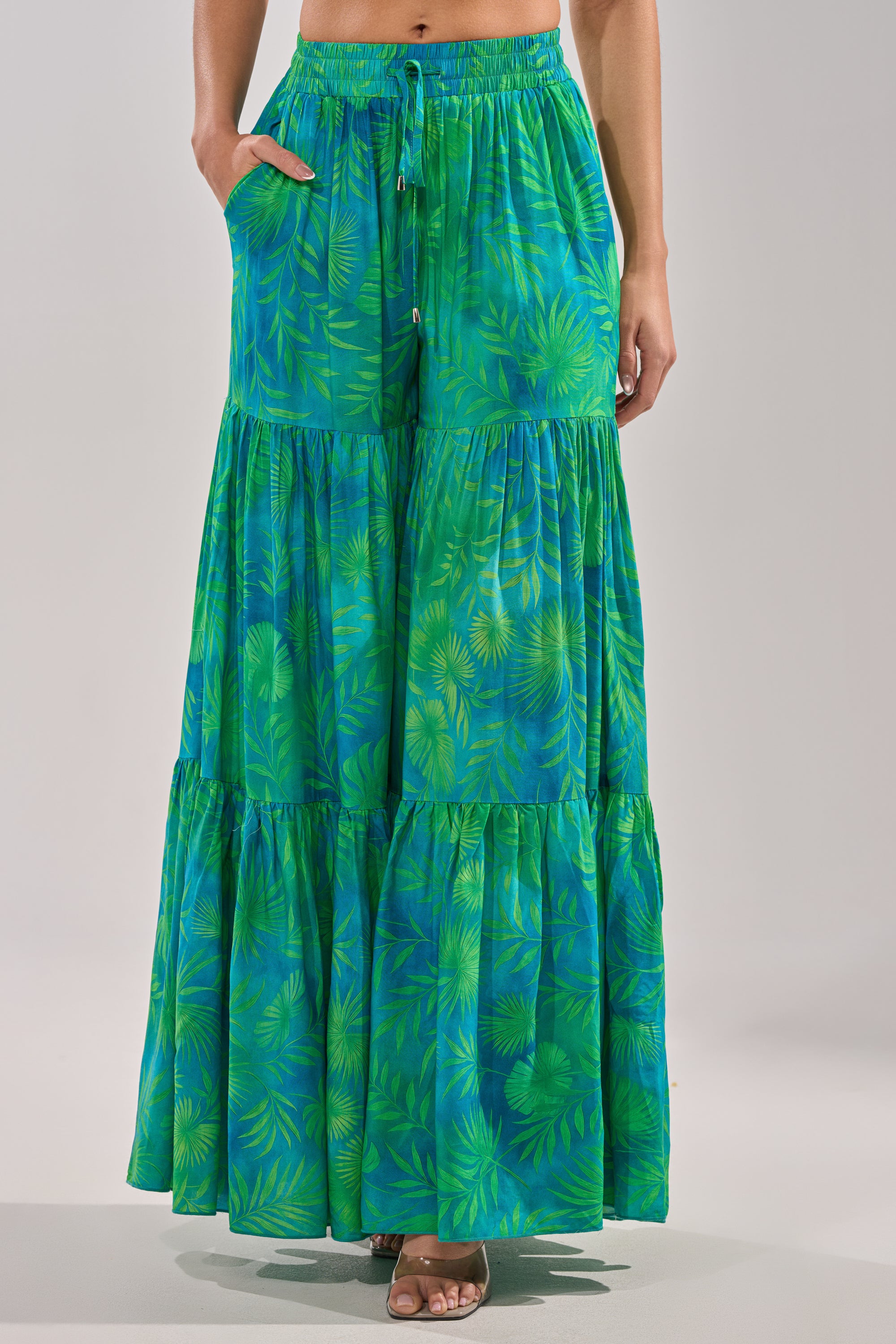 A person wears the INTO THE GARDEN PALAZZO PANT IN GREEN—long, tiered pants with a leafy, tropical print, hands in pockets and open-toe heels. Only the lower half of the body is visible. 4