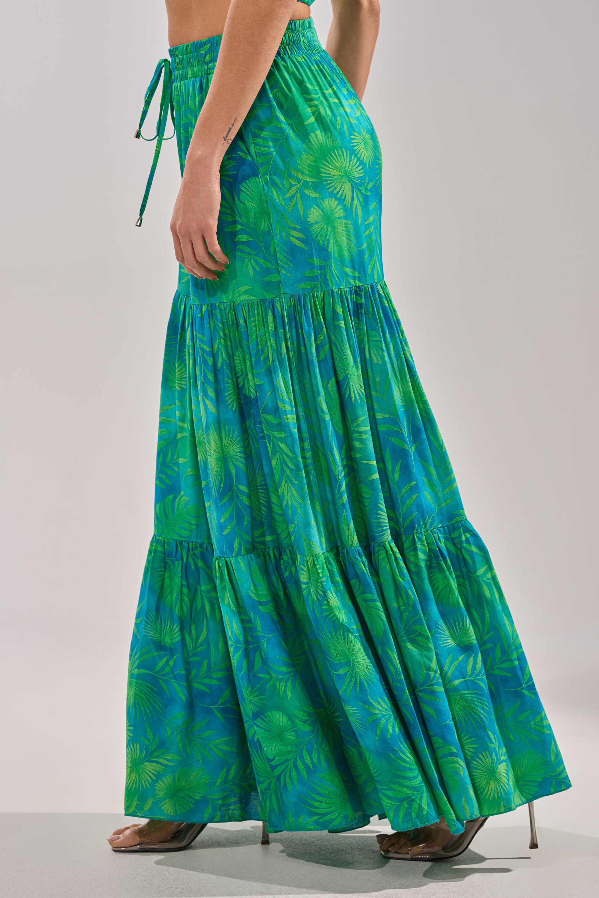 A woman models the INTO THE GARDEN PALAZZO PANT IN GREEN, featuring a tropical print and drawstring waist, paired with high-heeled sandals—evoking the effortless style of resort evening wear against a plain background. 5