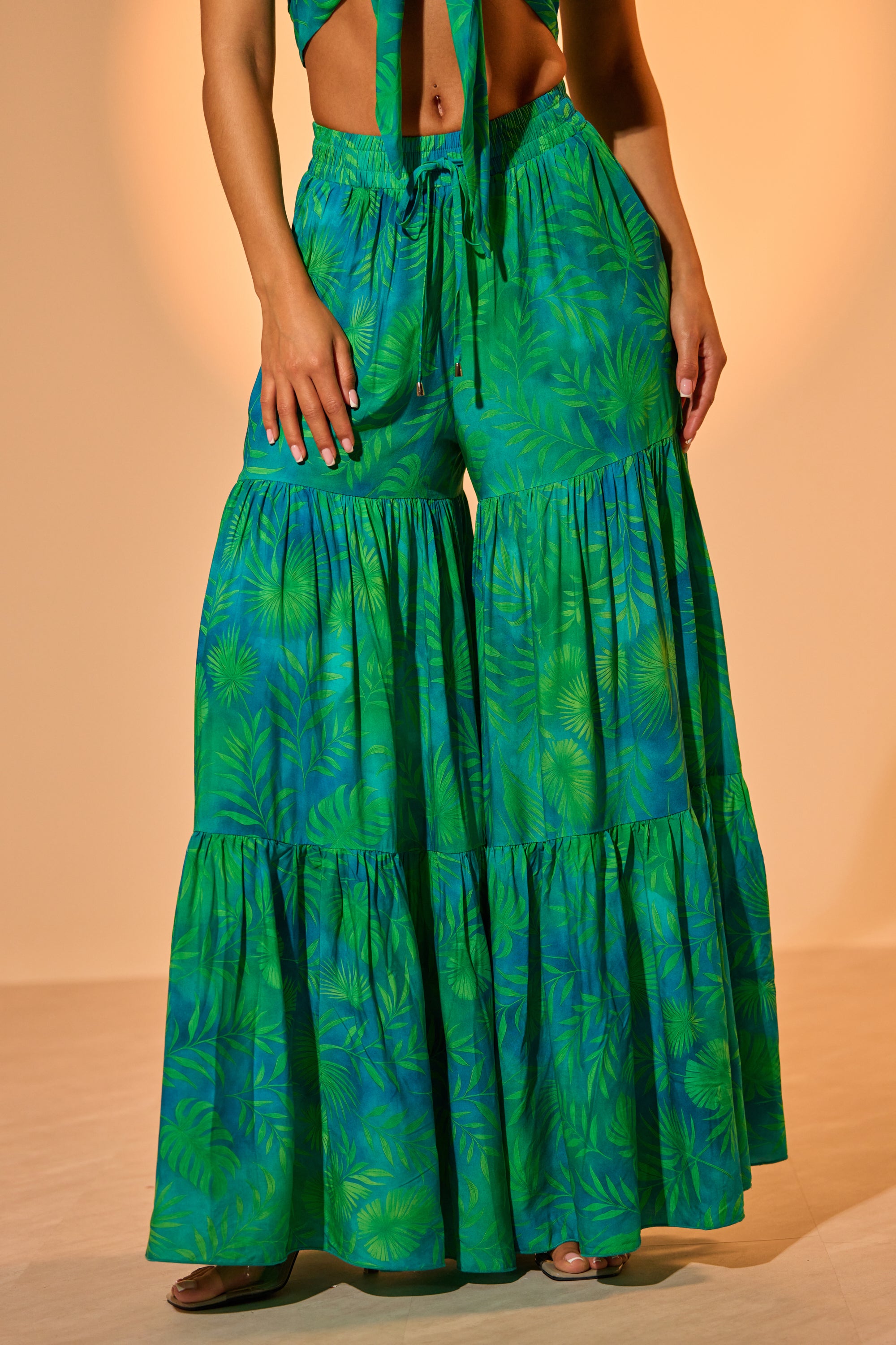 A woman stands barefoot in the INTO THE GARDEN PALAZZO PANT IN GREEN, featuring a high-waisted, wide-leg tropical leaf print. Her hands rest on a tier against a warm beige background, creating a relaxed resort evening vibe. 2
