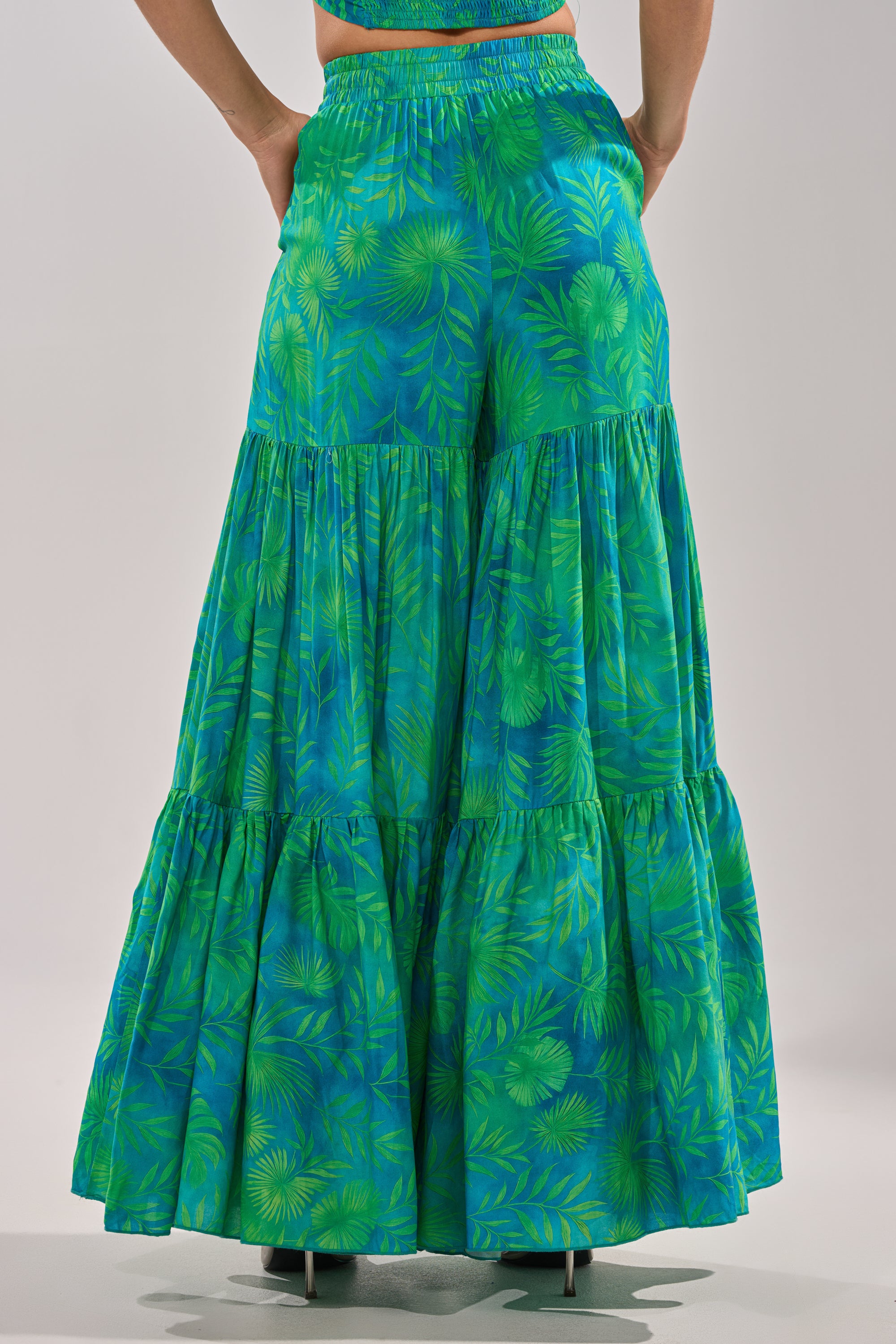 A person models the INTO THE GARDEN PALAZZO PANT IN GREEN, featuring a green and blue leaf print, wide legs, flowing fabric, and an elastic waistband. Photographed from the back against a plain background. 6