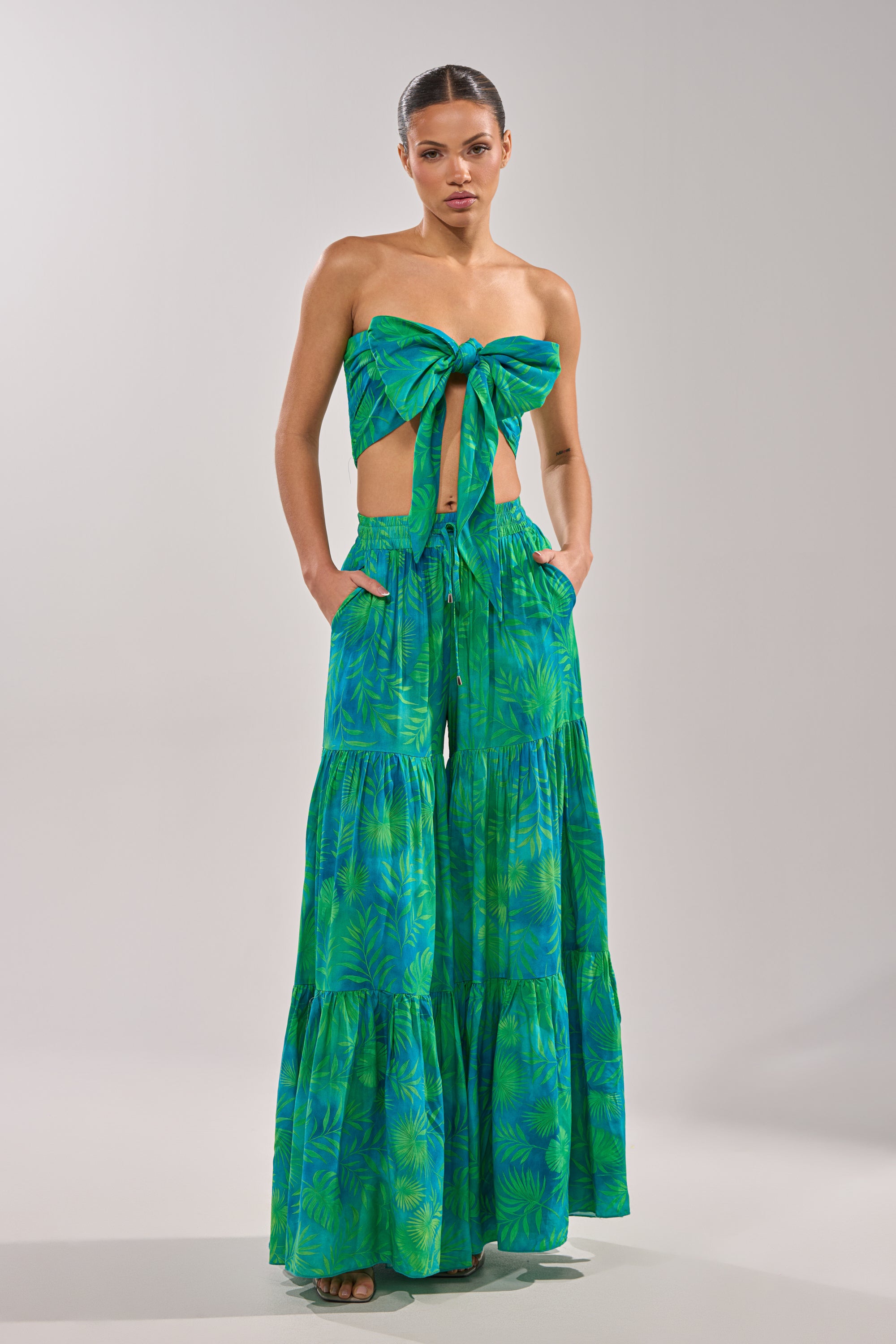 A woman poses with her hands in her pockets wearing the INTO THE GARDEN PALAZZO PANT IN GREEN, featuring a vibrant green tropical print and a matching strapless bow-tied top, with her hair pulled back against a plain background. 7