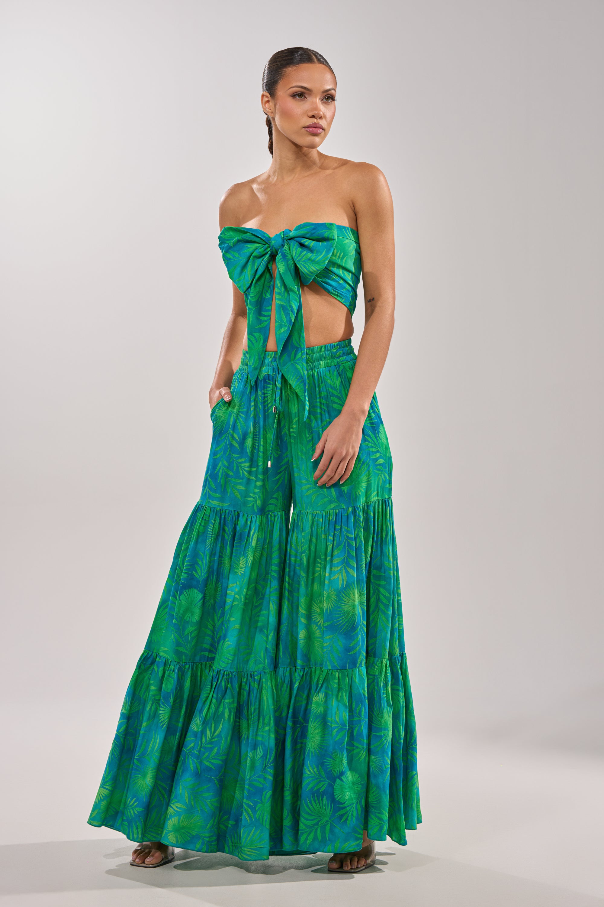 A woman wears the INTO THE GARDEN PALAZZO PANT IN GREEN: vibrant green, tropical-print, wide-leg pants paired with a matching strapless bow-tie crop top. Her hair is pulled back and her hands are in her pockets. 9