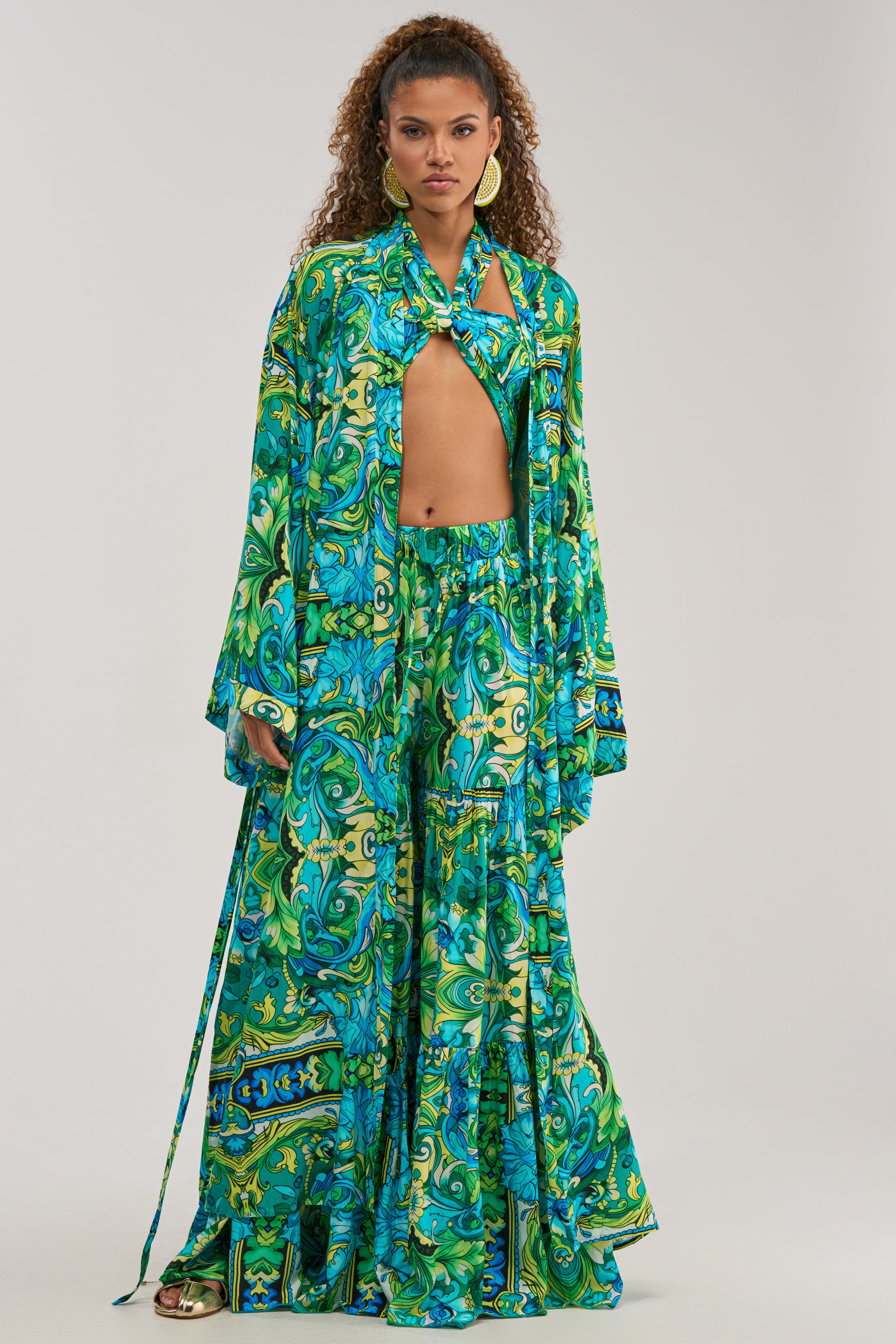 Draped in the INTO THE GARDEN DUSTER IN GREEN MULTI, this effortlessly chic summer ensemble features a flowing, botanical-patterned duster worn over a coordinating long skirt and styled with a scarf around the neck. Gold earrings and sandals complete the layered look.