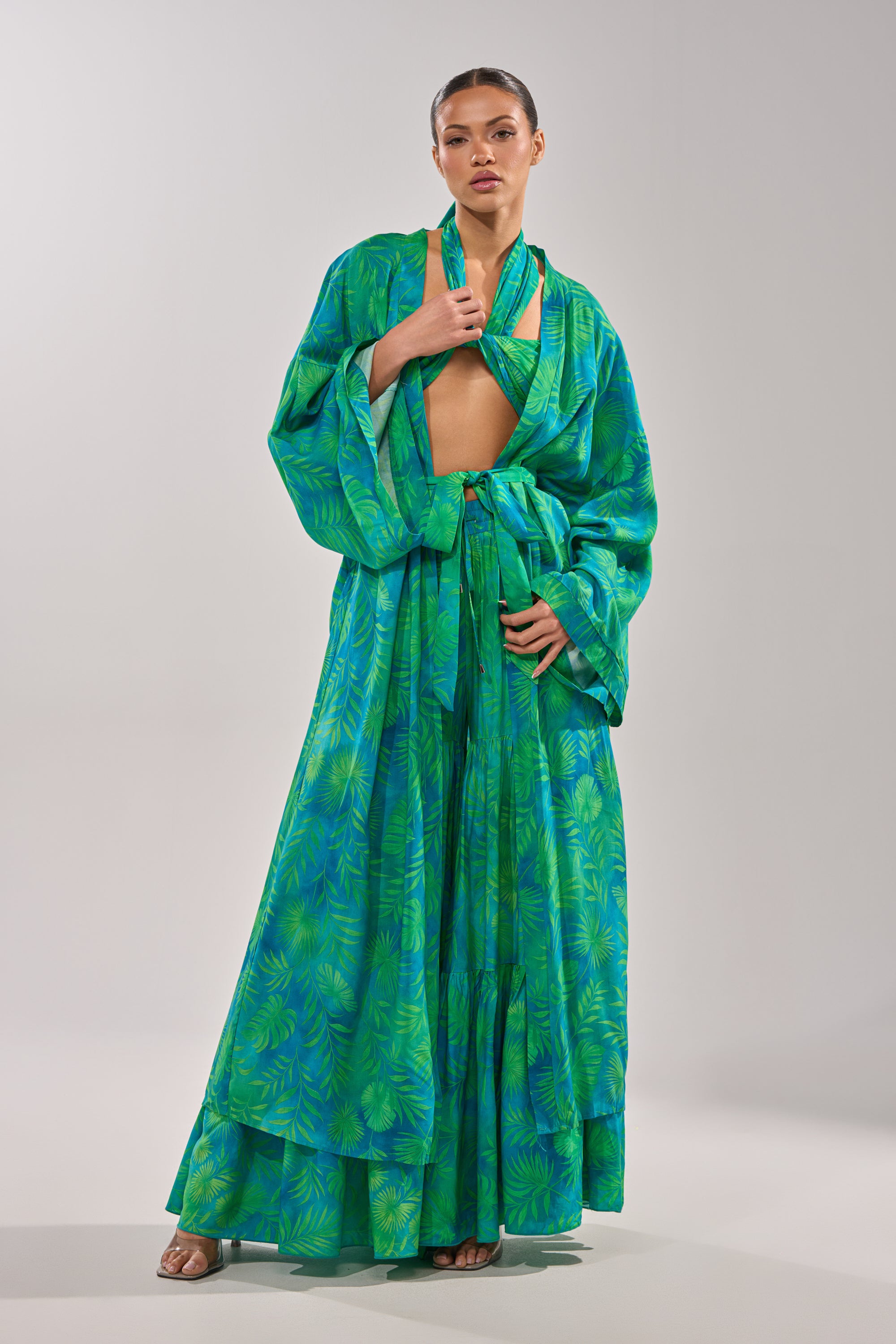 A woman models the INTO THE GARDEN DUSTER IN GREEN, a long, kimono-style dress with wide sleeves and a sash belt, showcasing its vibrant tropical print against a light gray background. 2