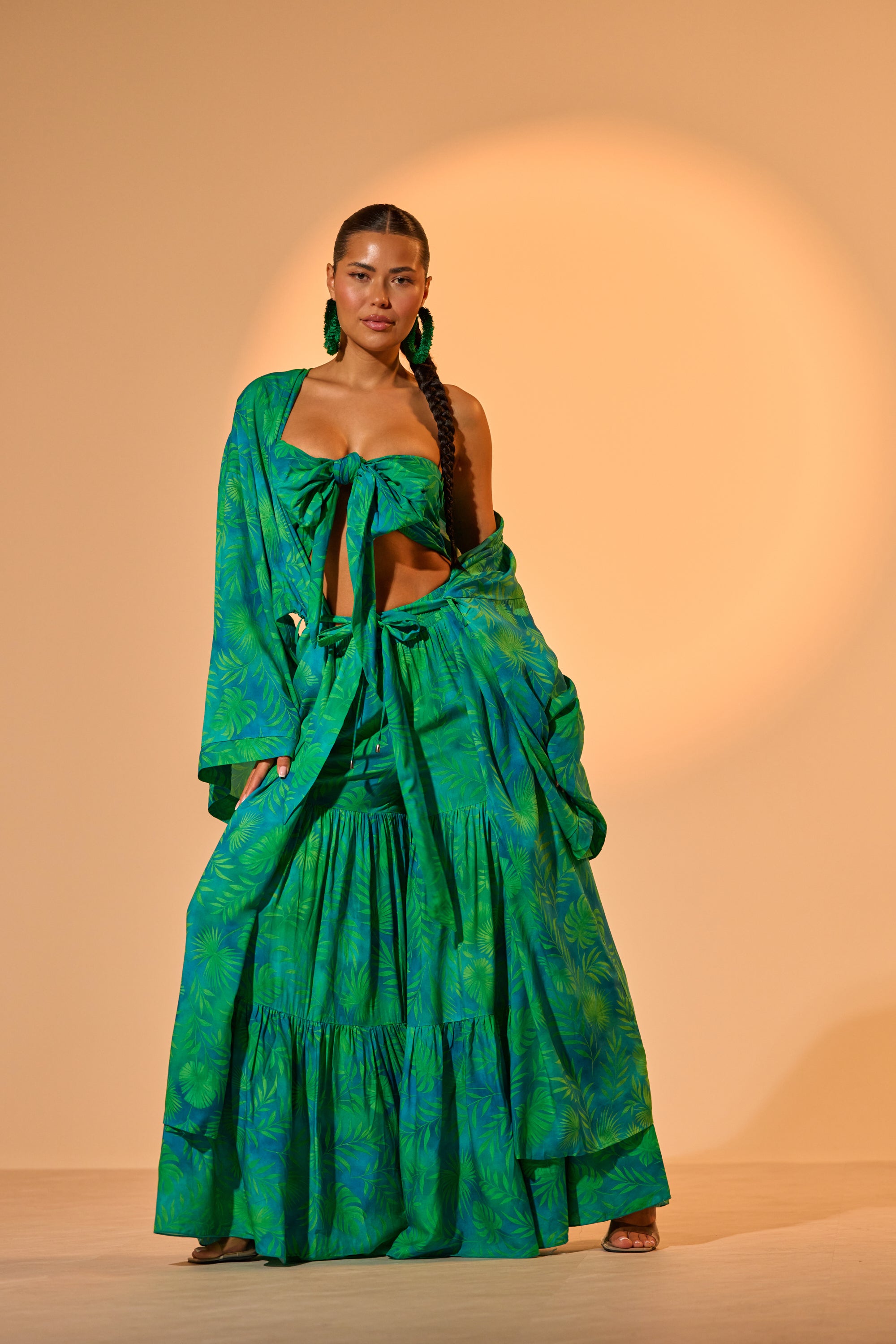 A woman stands confidently in the INTO THE GARDEN DUSTER IN GREEN, styled with long braids and green statement earrings, against a warm beige background—a chic vacation-ready look. 0