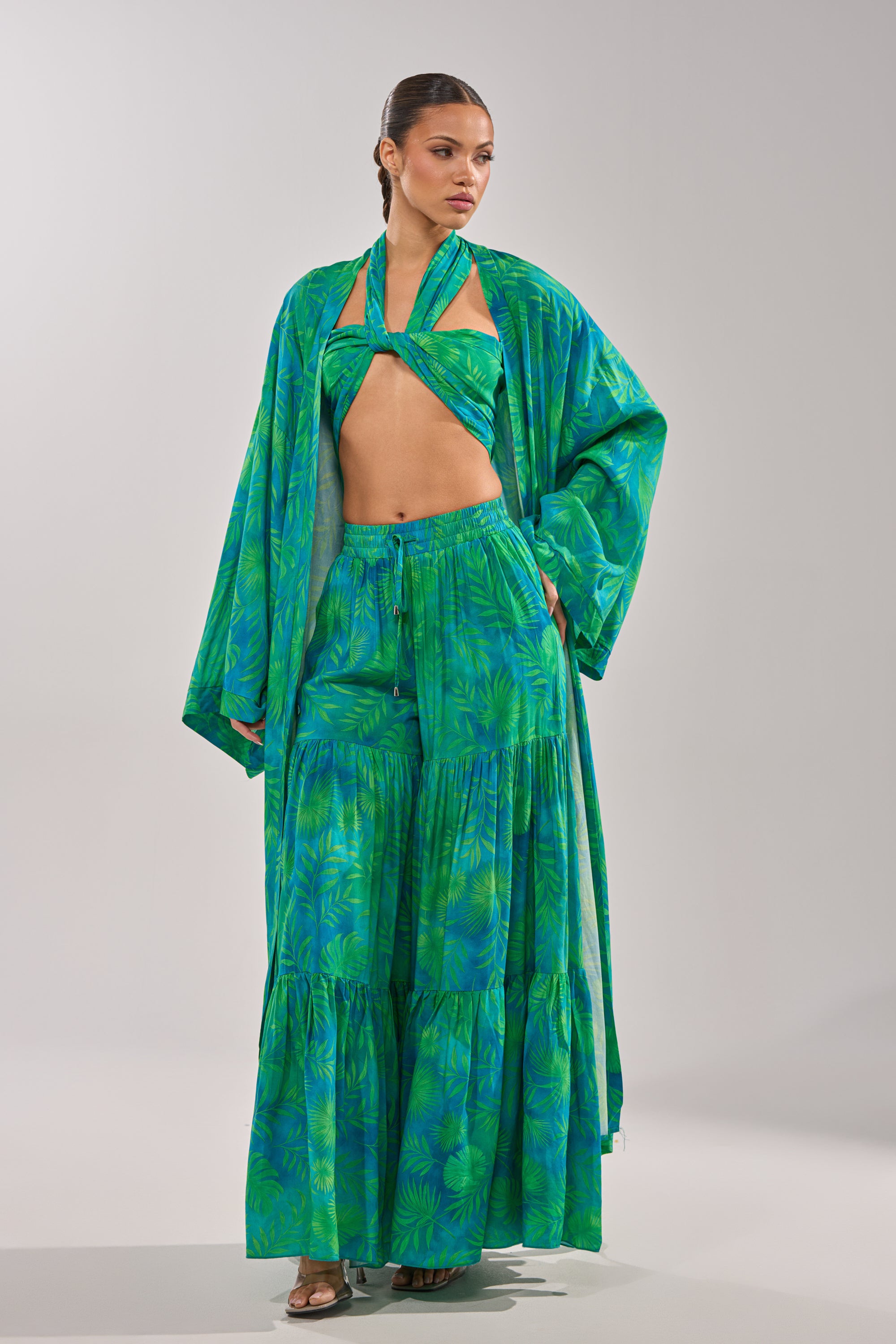 A woman wears the INTO THE GARDEN DUSTER IN GREEN, a vibrant green botanical print set with a halter top, long tiered skirt, and matching duster, standing confidently against a plain light gray background. 3