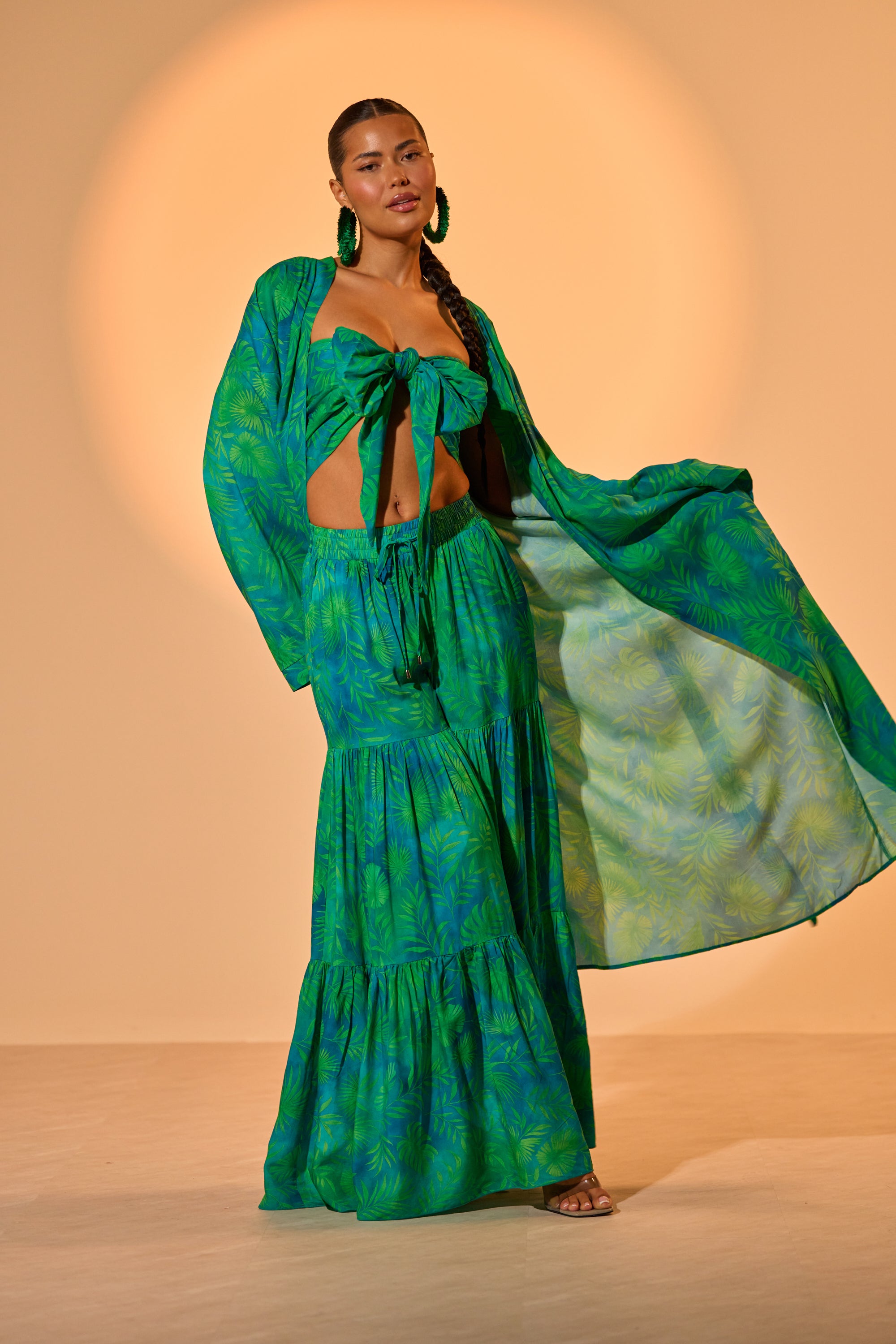A woman models the INTO THE GARDEN DUSTER IN GREEN, a vibrant tropical print set with a crop top, long flowy skirt, and matching duster, posed on a neutral background with warm lighting. 1