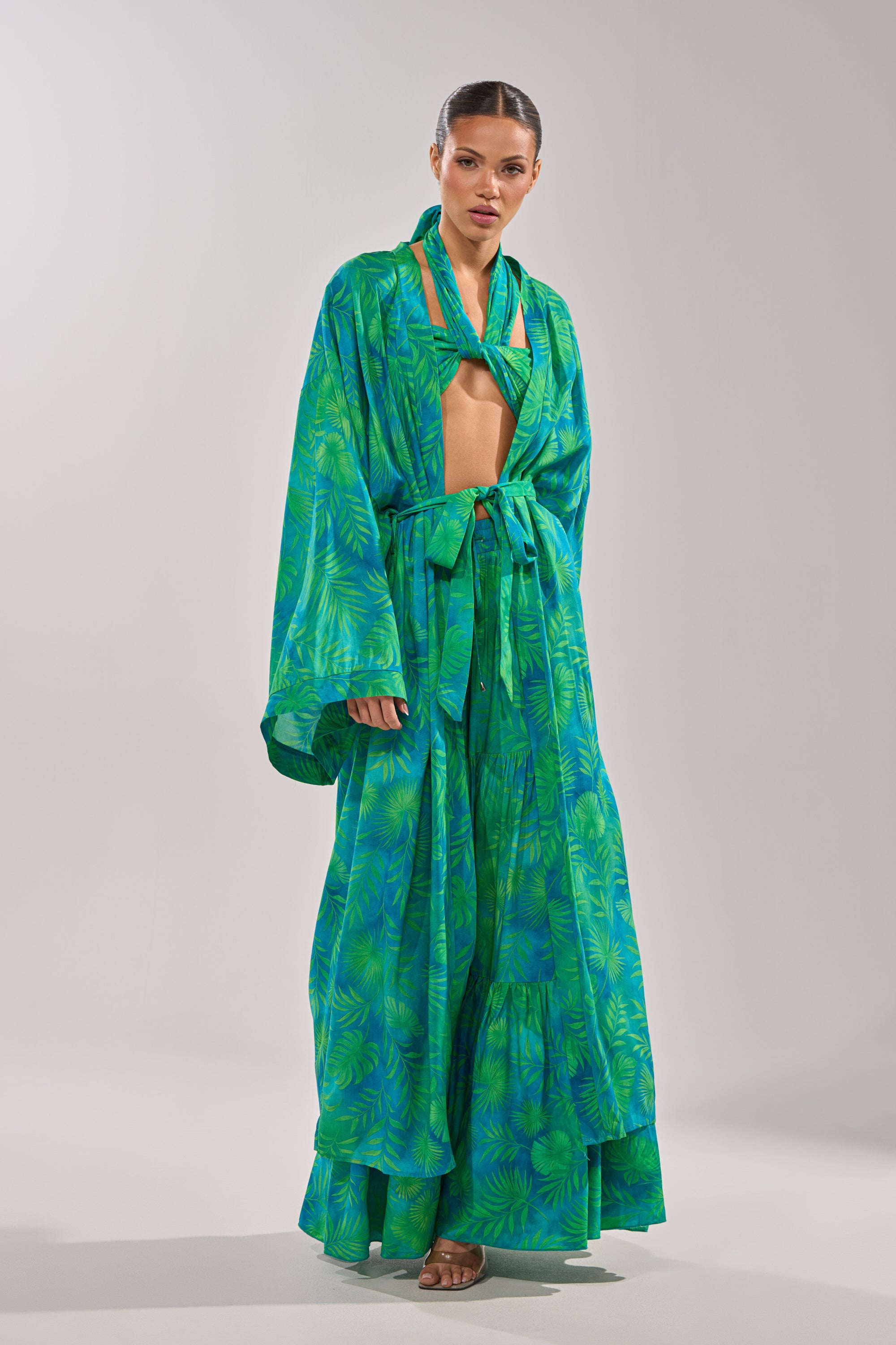 A woman wears the INTO THE GARDEN DUSTER IN GREEN, a vibrant green and blue botanical vacation set with a tropical print duster, wide sleeves, tie belt, matching flowing skirt, bikini-style top, slicked-back hair, and neutral sandals. 5