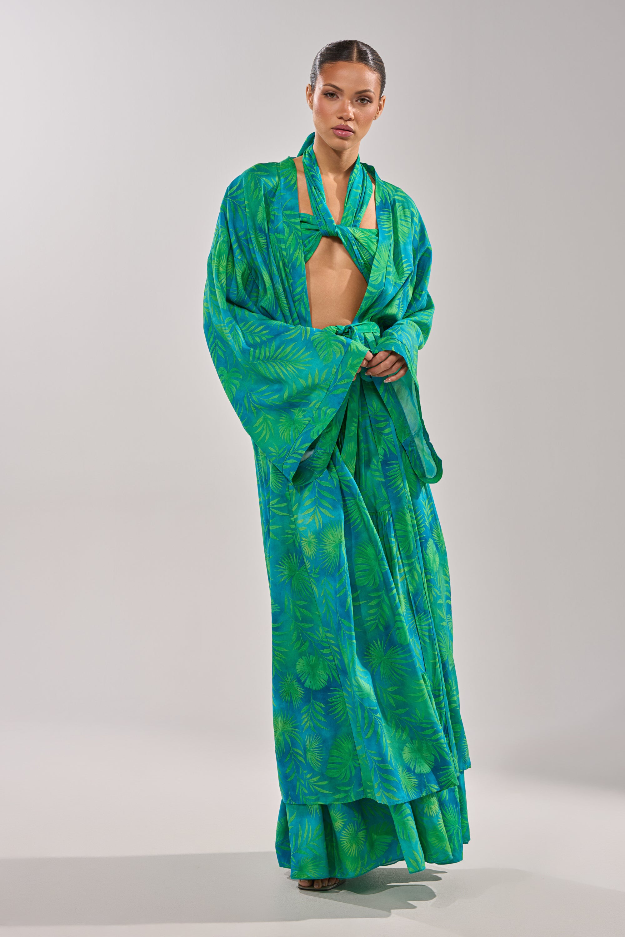 A woman models the INTO THE GARDEN DUSTER IN GREEN, featuring a vibrant green and blue botanical pattern, wide sleeves, and a wrapped neckline, against a plain light background. 6