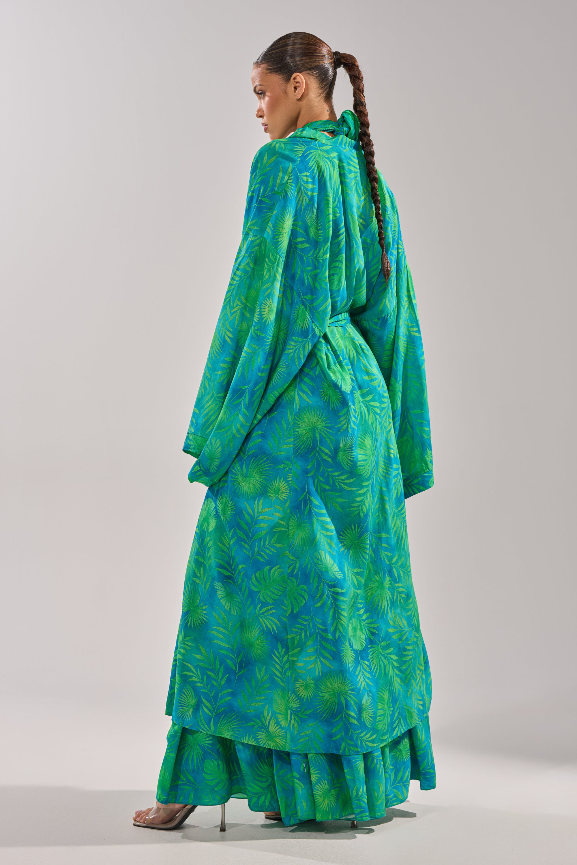 A person stands sideways wearing the INTO THE GARDEN DUSTER IN GREEN, a long, flowing duster with a green and blue botanical pattern, wide sleeves, and layered tiers. Their hair is styled in a sleek braided ponytail and they wear clear high-heeled shoes. 7
