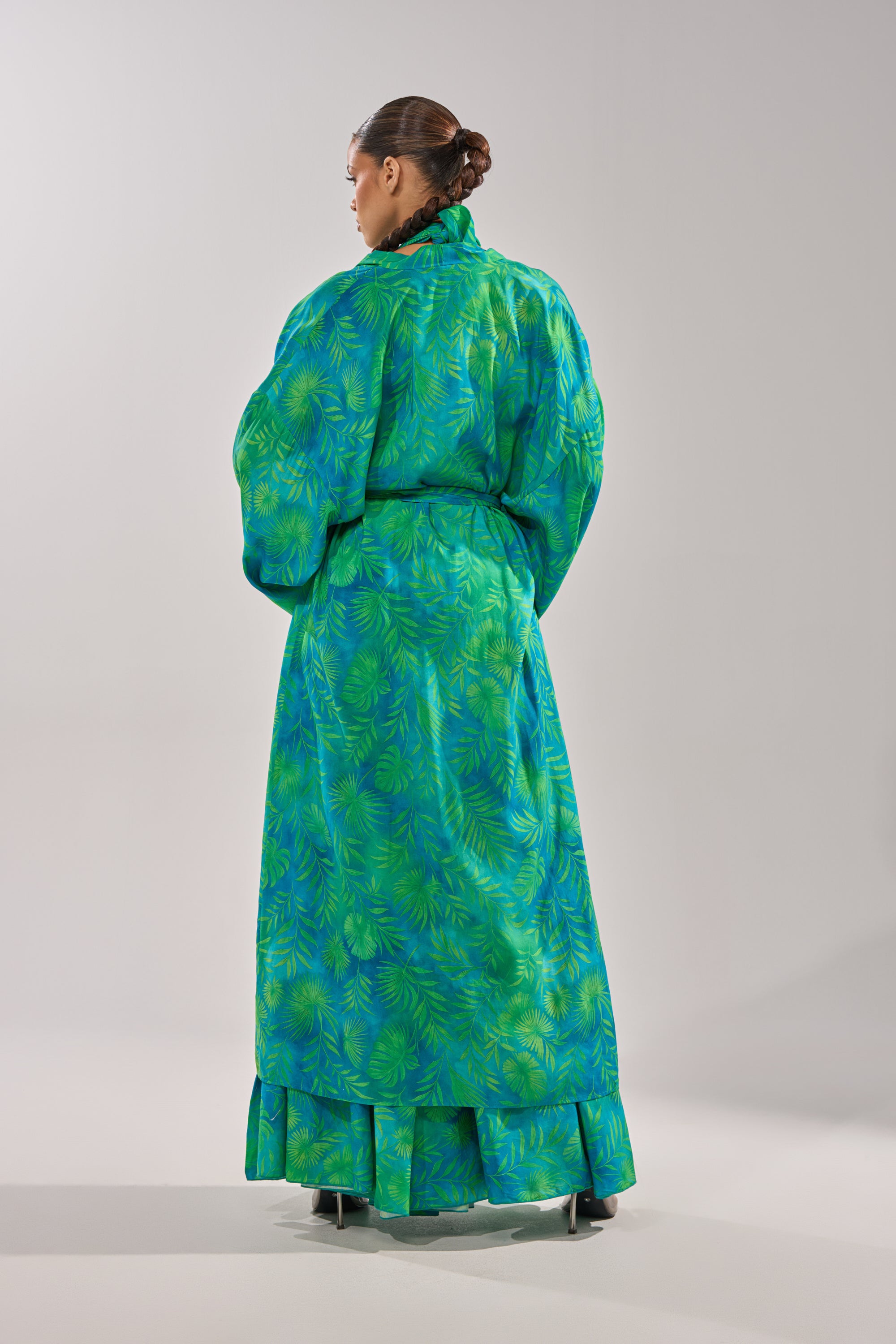 A person stands facing away, wearing the INTO THE GARDEN DUSTER IN GREEN—a long, belted dress with a botanical pattern in bright green and blue, featuring loose sleeves and a ruffled hem. Hair is styled in a bun against a plain light gray background. 8