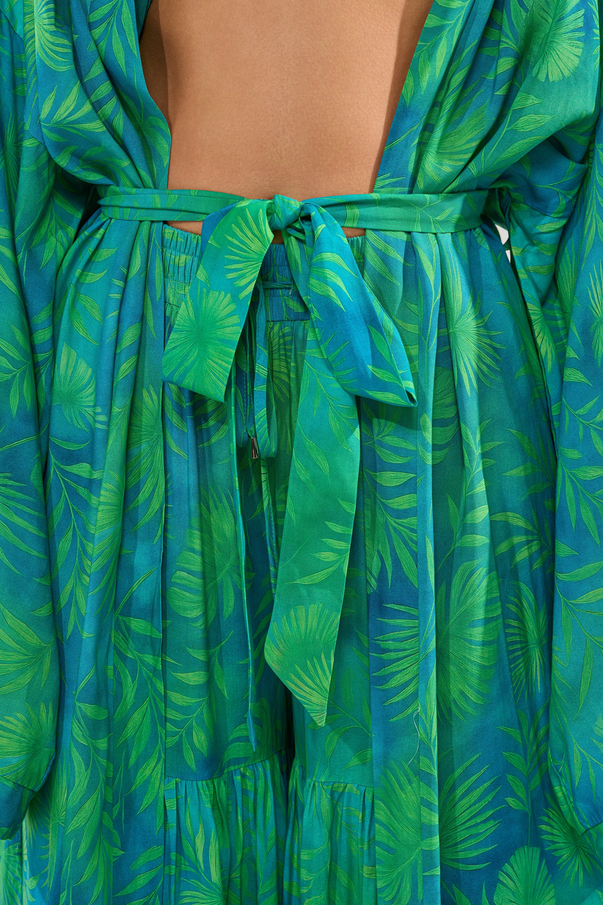 A close-up of a person wearing the INTO THE GARDEN DUSTER IN GREEN, featuring a sheer blue and green tropical print with a matching fabric belt tied at the waist and their bare back partially visible above the belt. 9