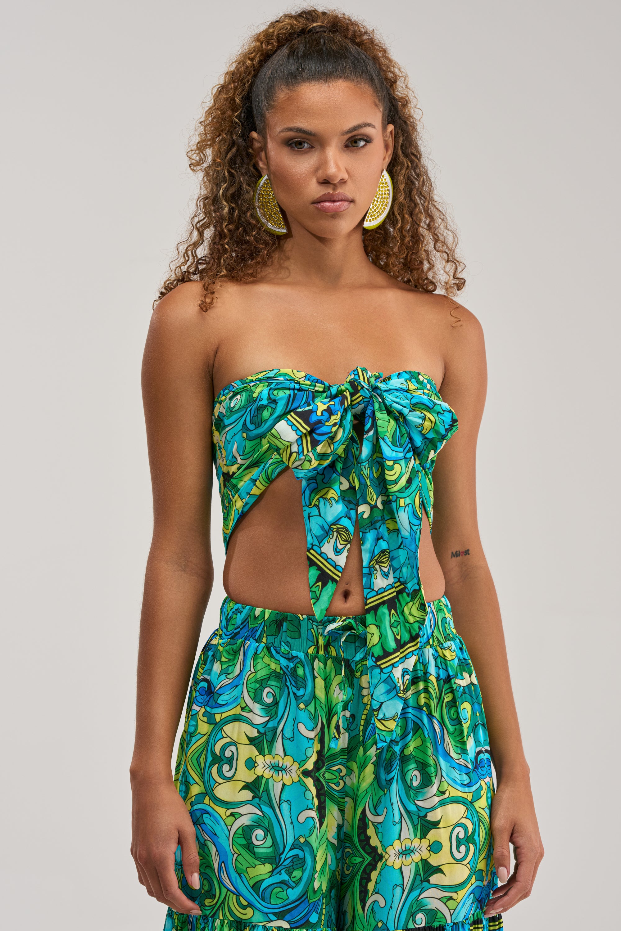 A woman wearing the INTO THE GARDEN BRALETTE IN GREEN MULTI—a vibrant blue and green patterned tube top with a front bow—paired with matching pants, perfect for festival clothing, stands against a plain background.