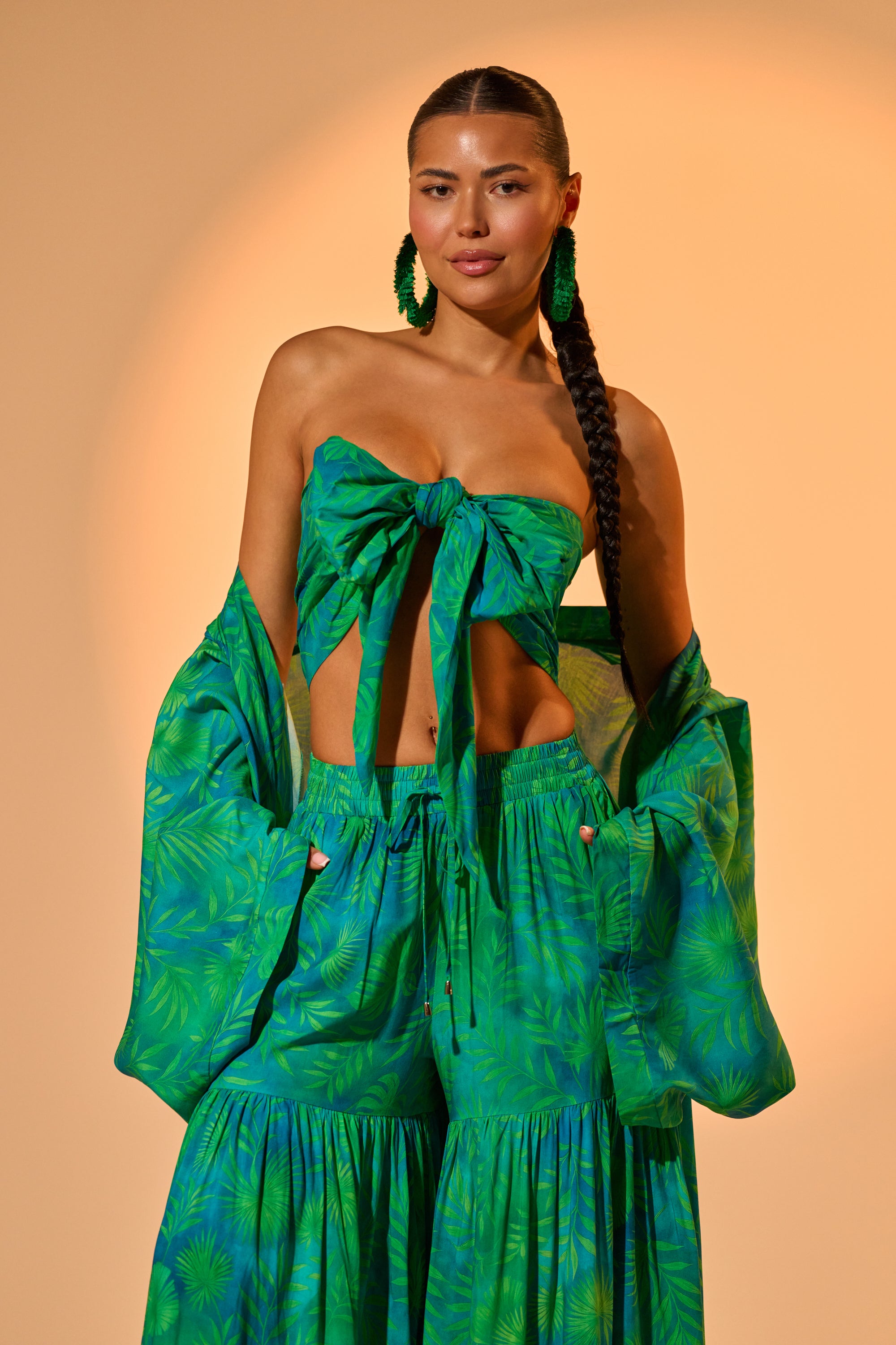A woman with a long braid wears the INTO THE GARDEN BRALETTE IN GREEN, styled with a matching tropical cover-up and flowy pants, and poses confidently against a beige background. 0