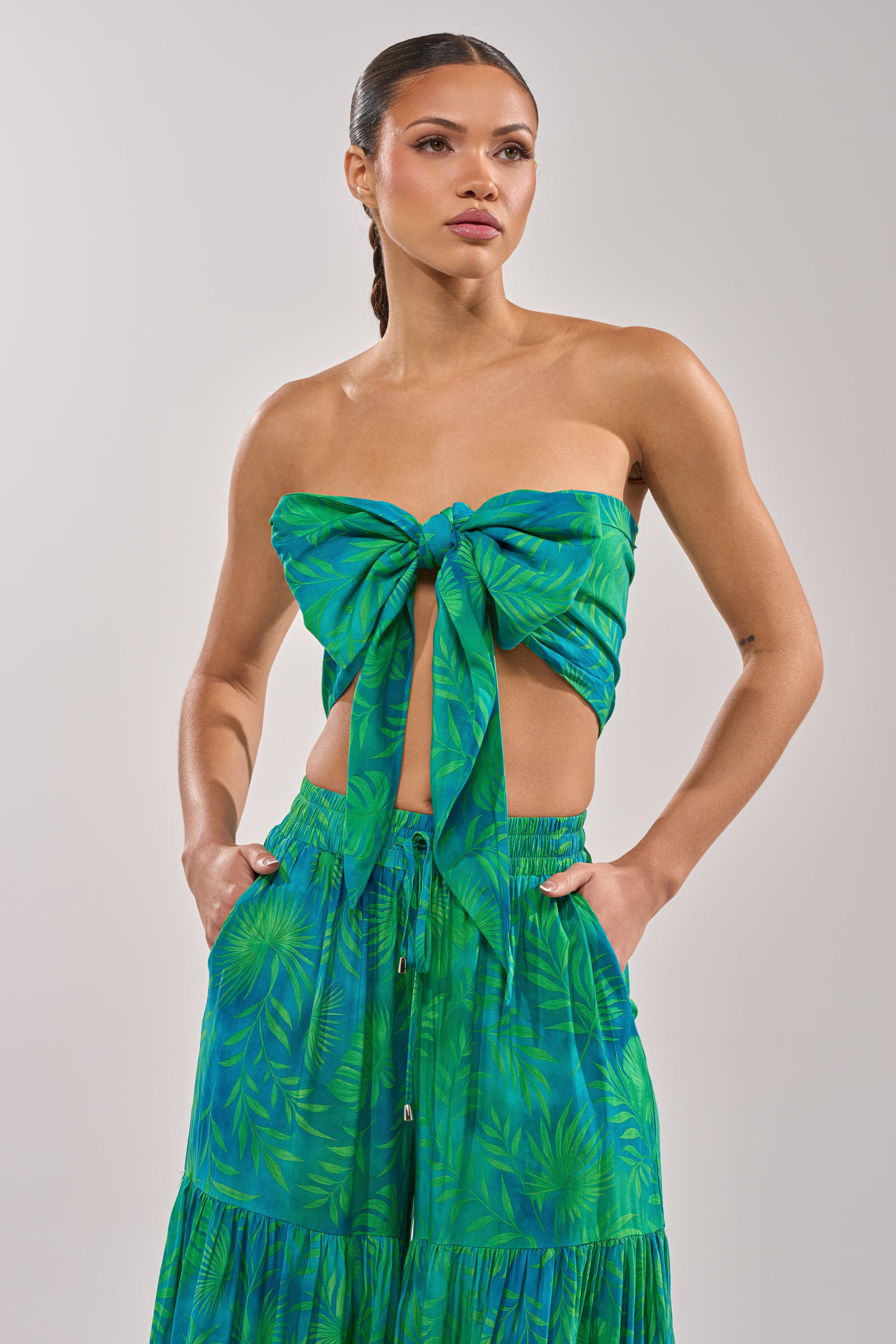 A woman wears the INTO THE GARDEN BRALETTE IN GREEN, a halter-style green and blue tropical print top tied in a bow, paired with a matching skirt, standing with hands in pockets against a plain light background. 3