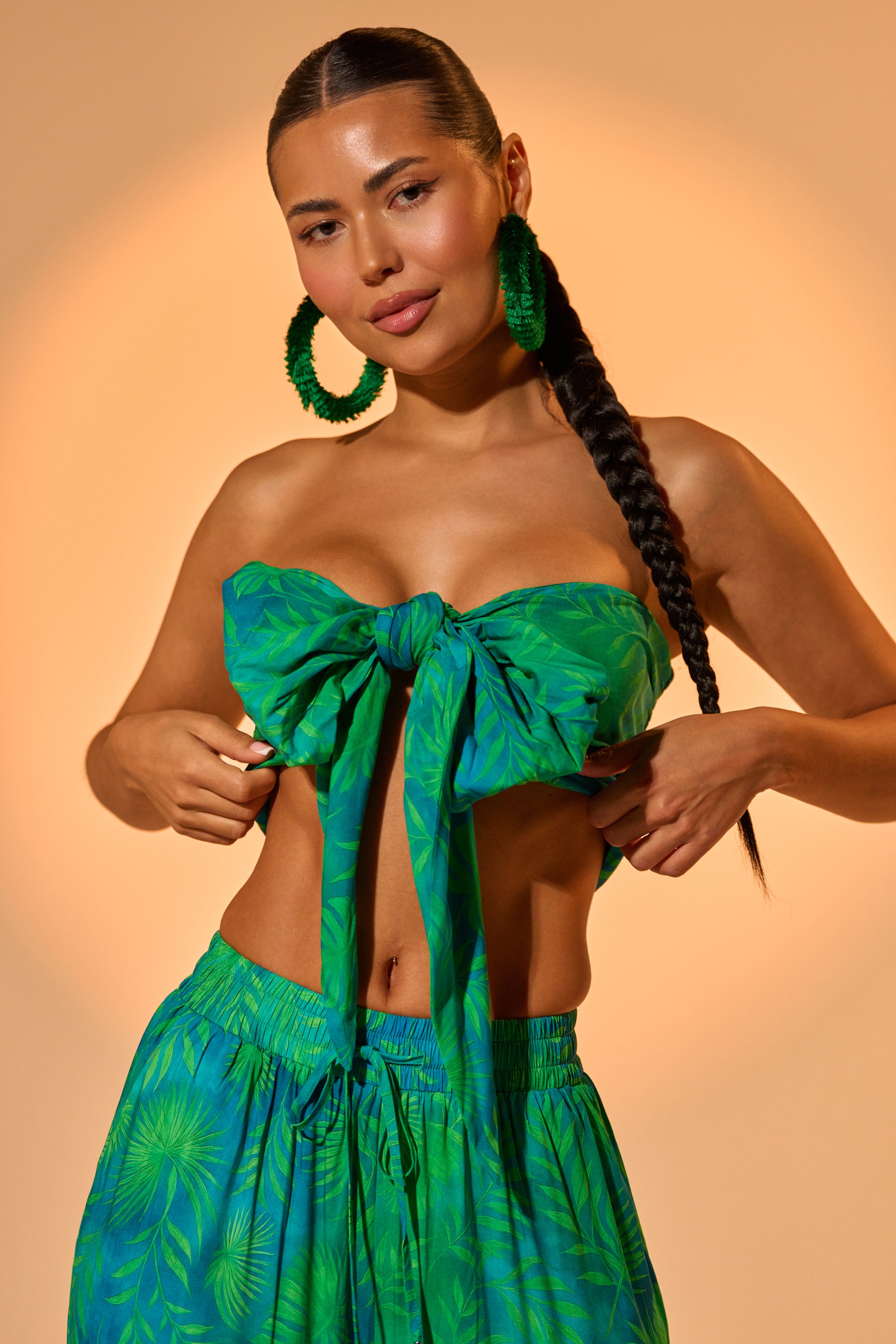 A woman with a long braid wears the INTO THE GARDEN BRALETTE IN GREEN and matching skirt, styled as chic resort wear. She accessorizes with large green hoop earrings and poses before a warm, peach-toned background. 1