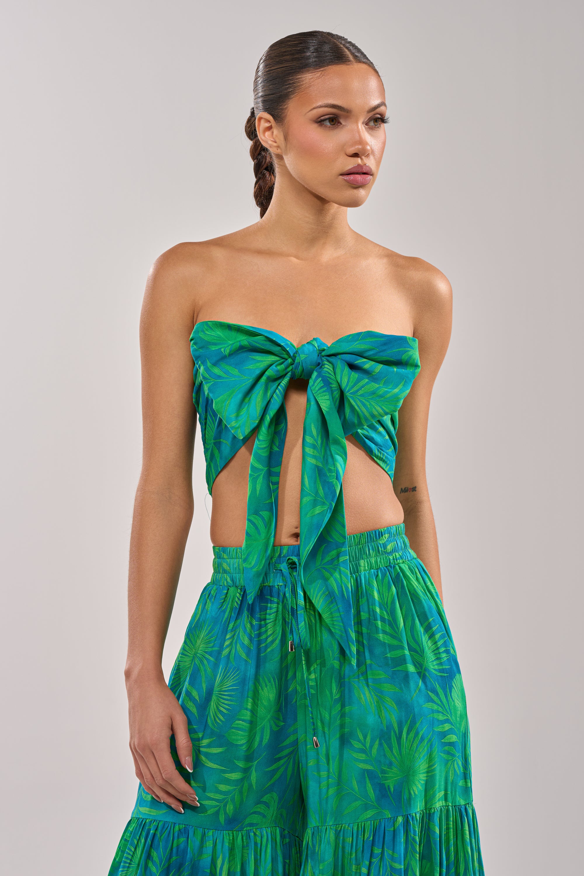 A woman models the INTO THE GARDEN BRALETTE IN GREEN, a strapless tropical print top tied in front with a matching high-waisted skirt—ideal for resort wear. 5