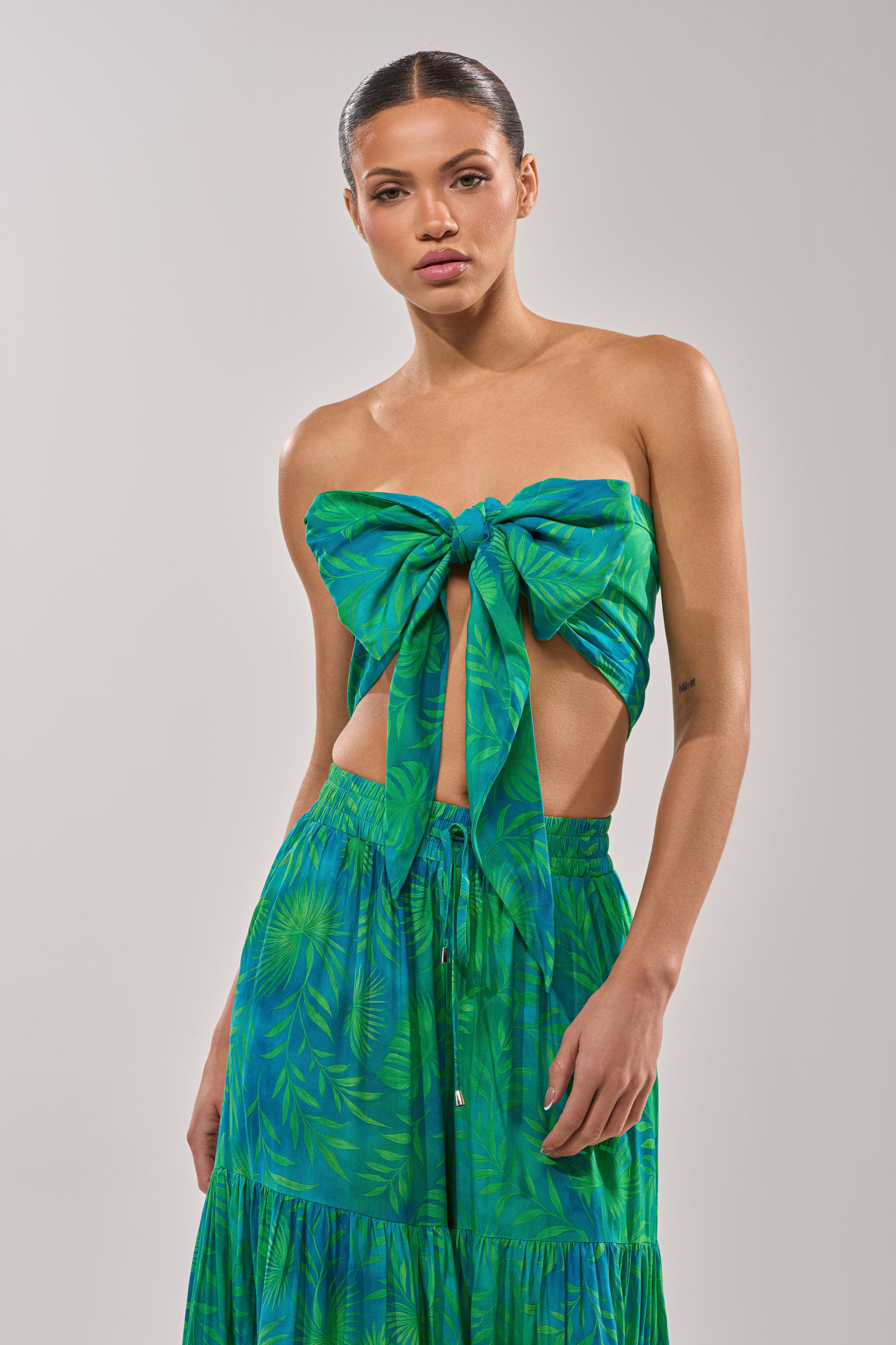 A woman wears the INTO THE GARDEN BRALETTE IN GREEN, a strapless green and blue tropical print top tied in a large bow, with a matching skirt, standing against a plain light gray background. 6