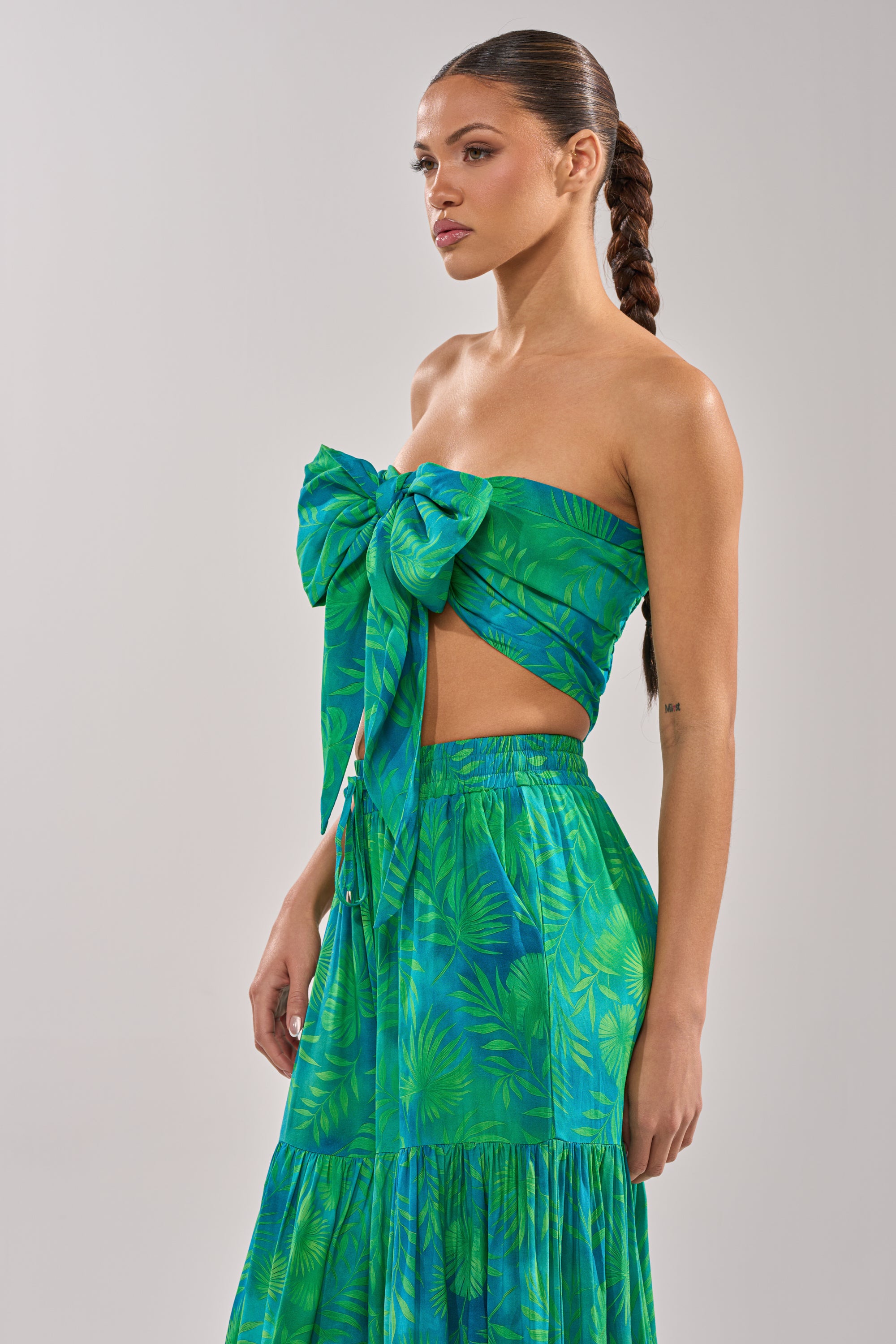 A woman with a long braided ponytail wears the INTO THE GARDEN BRALETTE IN GREEN, a tropical print crop top with a large front bow and matching high-waisted skirt, perfect for resort wear, against a plain light gray background. 7