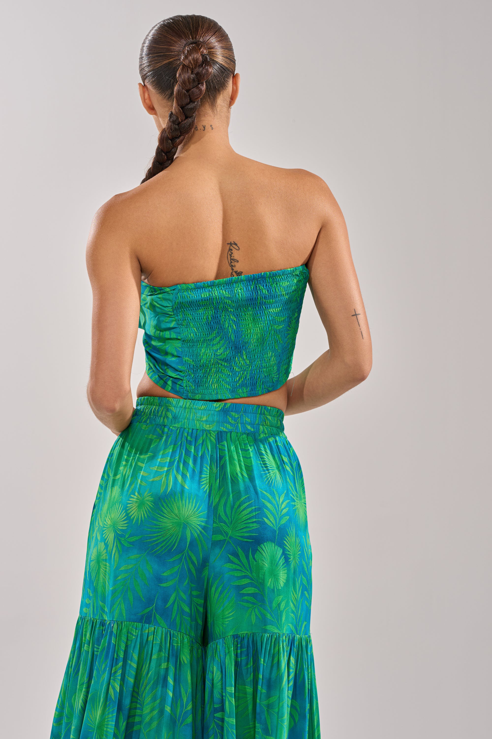 A woman with a long braided ponytail and tattoos wears the INTO THE GARDEN BRALETTE IN GREEN, a halter-style tropical print top, paired with a matching skirt for a chic resort-ready look. 8