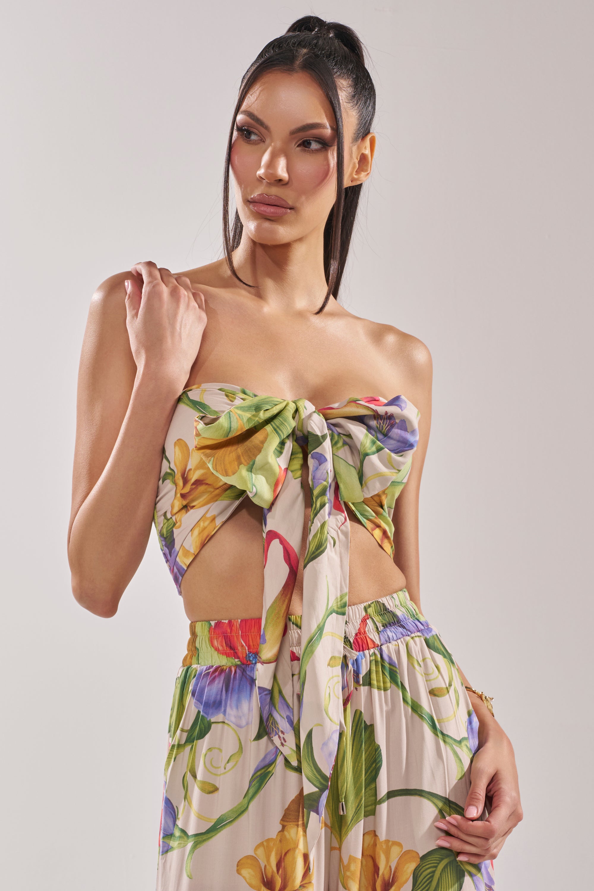 A woman with long dark hair in a high ponytail wears the INTO THE GARDEN FLORAL BLOUSE, featuring a strapless neckline and paired with matching high-waisted pants. She poses with one hand on her shoulder against a plain background.
